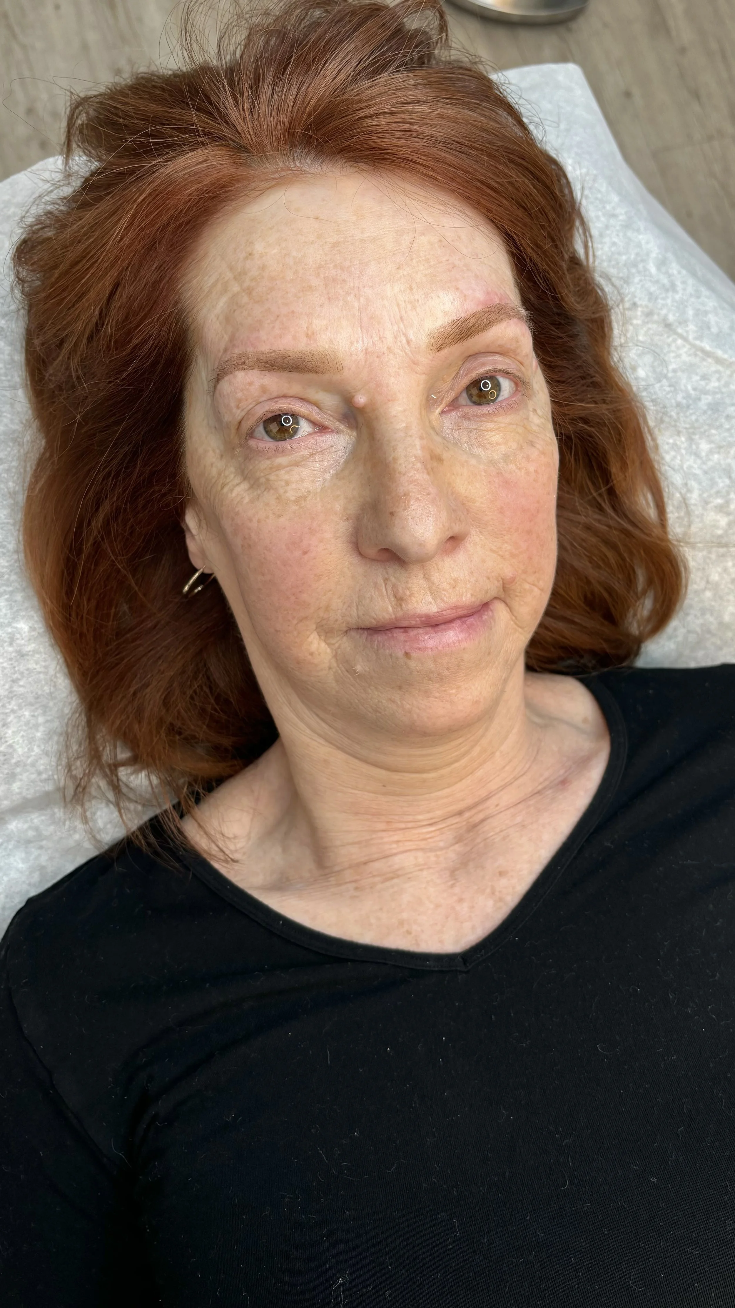 A woman with red hair lying down, showing her face with a small mole on her nose, detailed eyebrows, and slight makeup, in a relaxed setting.