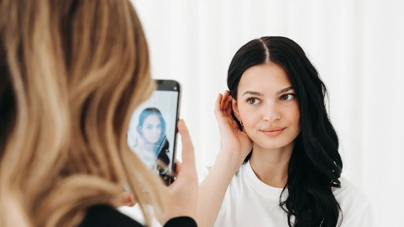 A woman with long, dark hair and a white shirt being photographed by another woman with blonde hair using a smartphone. The woman with dark hair is adjusting her earring and smiling softly.