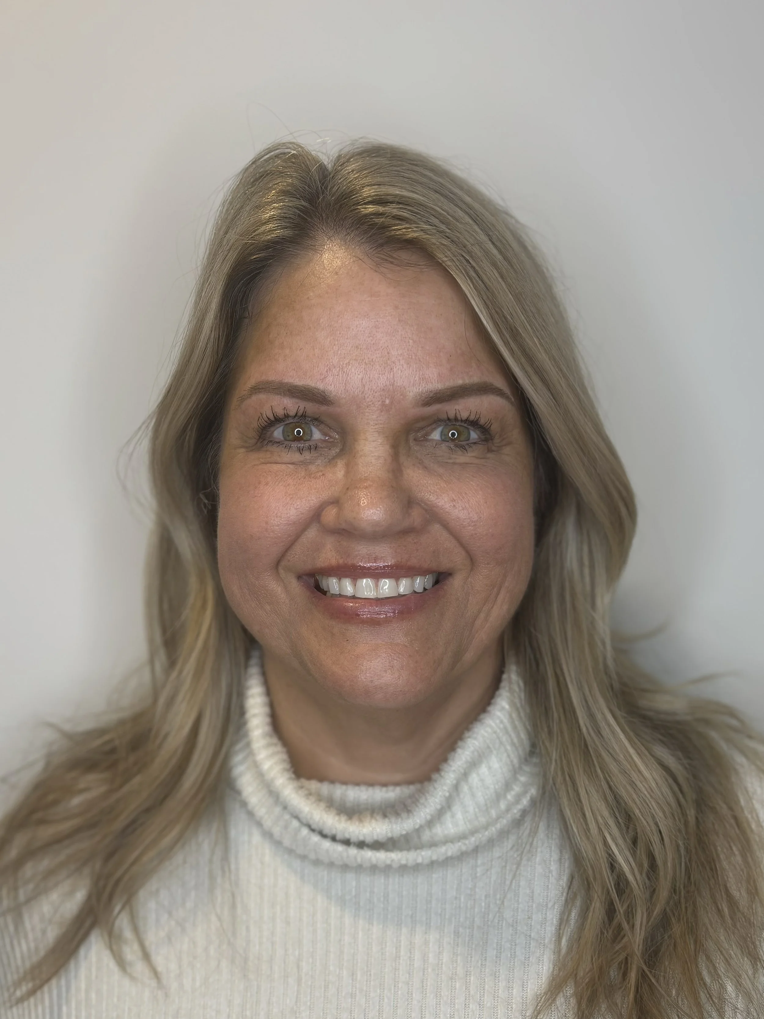 Close-up of a smiling woman with blonde hair and light-colored eyes, wearing a white turtleneck sweater, against a plain white background.