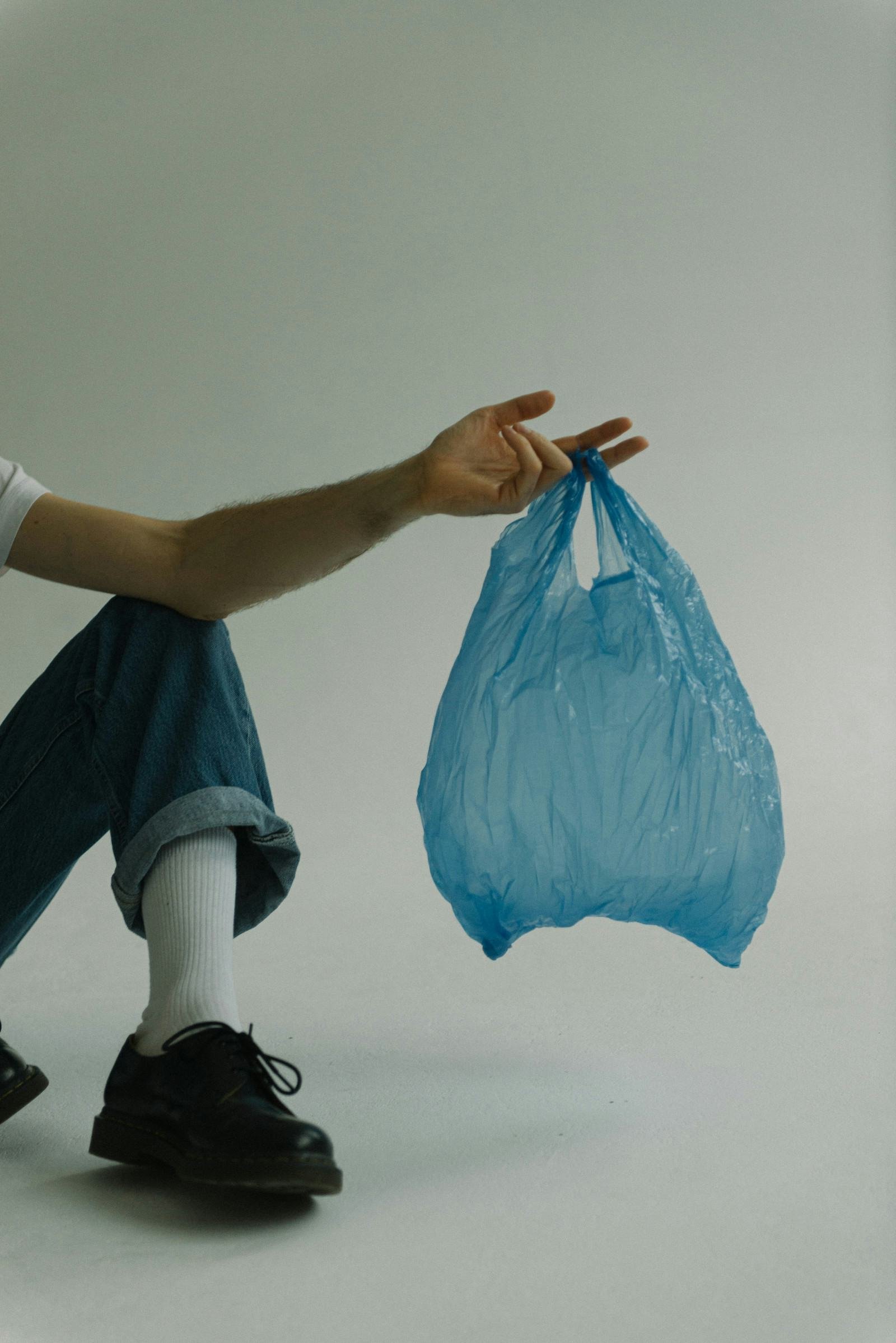 Person squatting and holding a blue plastic bag in mid-air with their right hand, against a plain wall.