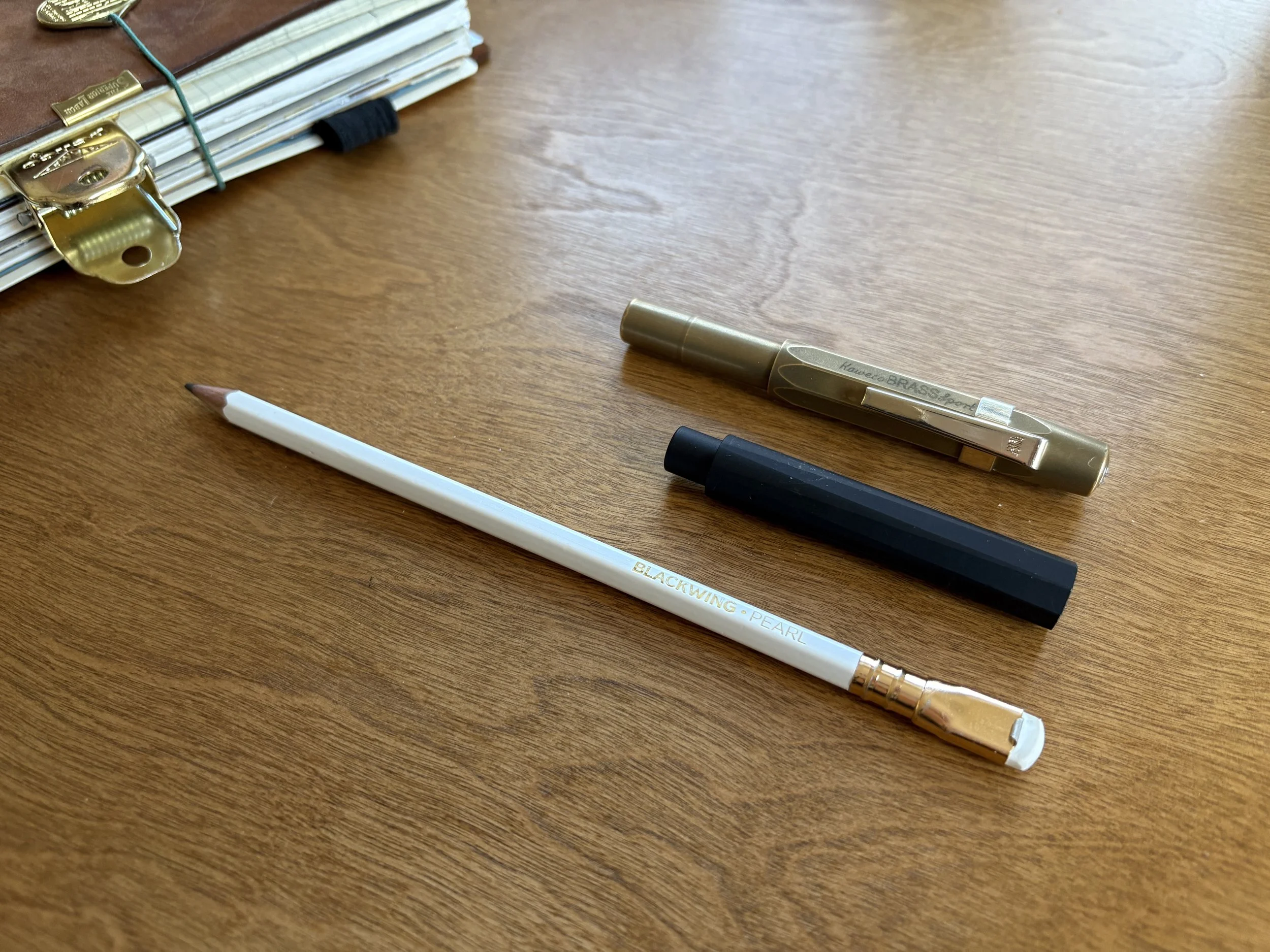 Minimalist art supplies on a wood desk - Blackwing pearl pencil, Black Tom's Studio Wren pen, Kaweco Brass Sport fountain pen, Traveler's notebook in background