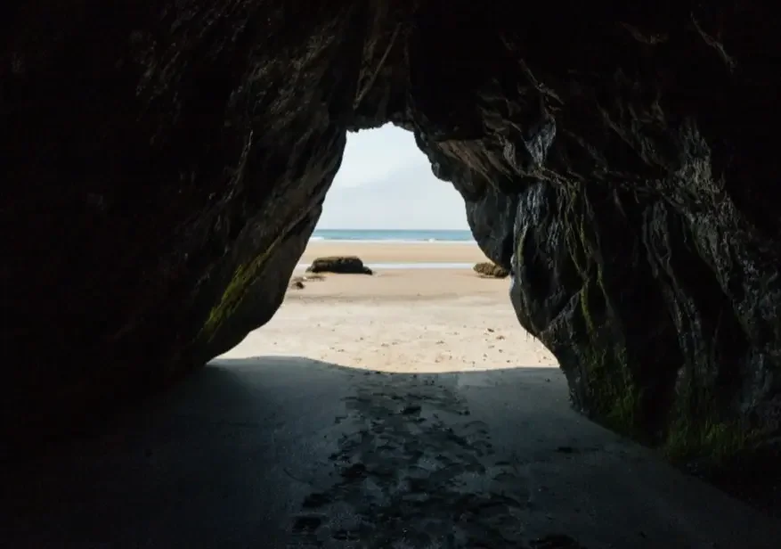 View through a rocky cave opening onto a sandy beach and ocean with clouds in the sky.