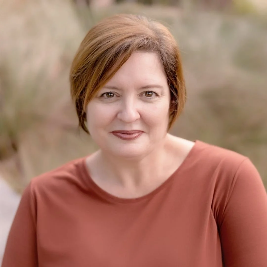 A woman with curly black hair smiling with her hand resting on her chin, wearing a white sleeveless top with embellishments, a gold watch, and earrings, against a neutral background.