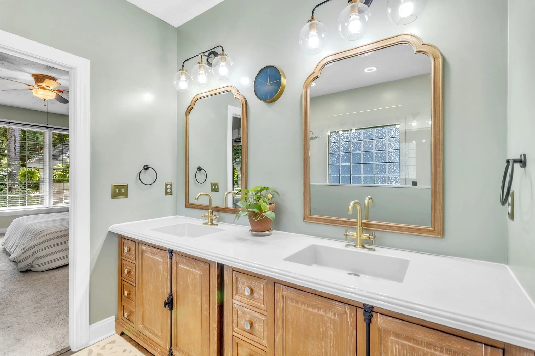 Bathroom with a double vanity, wooden cabinets, gold faucets, two large wooden-framed mirrors, a wall clock, a potted plant, and a light fixture with multiple bulbs. Visible bedroom with a ceiling fan and window in the background.