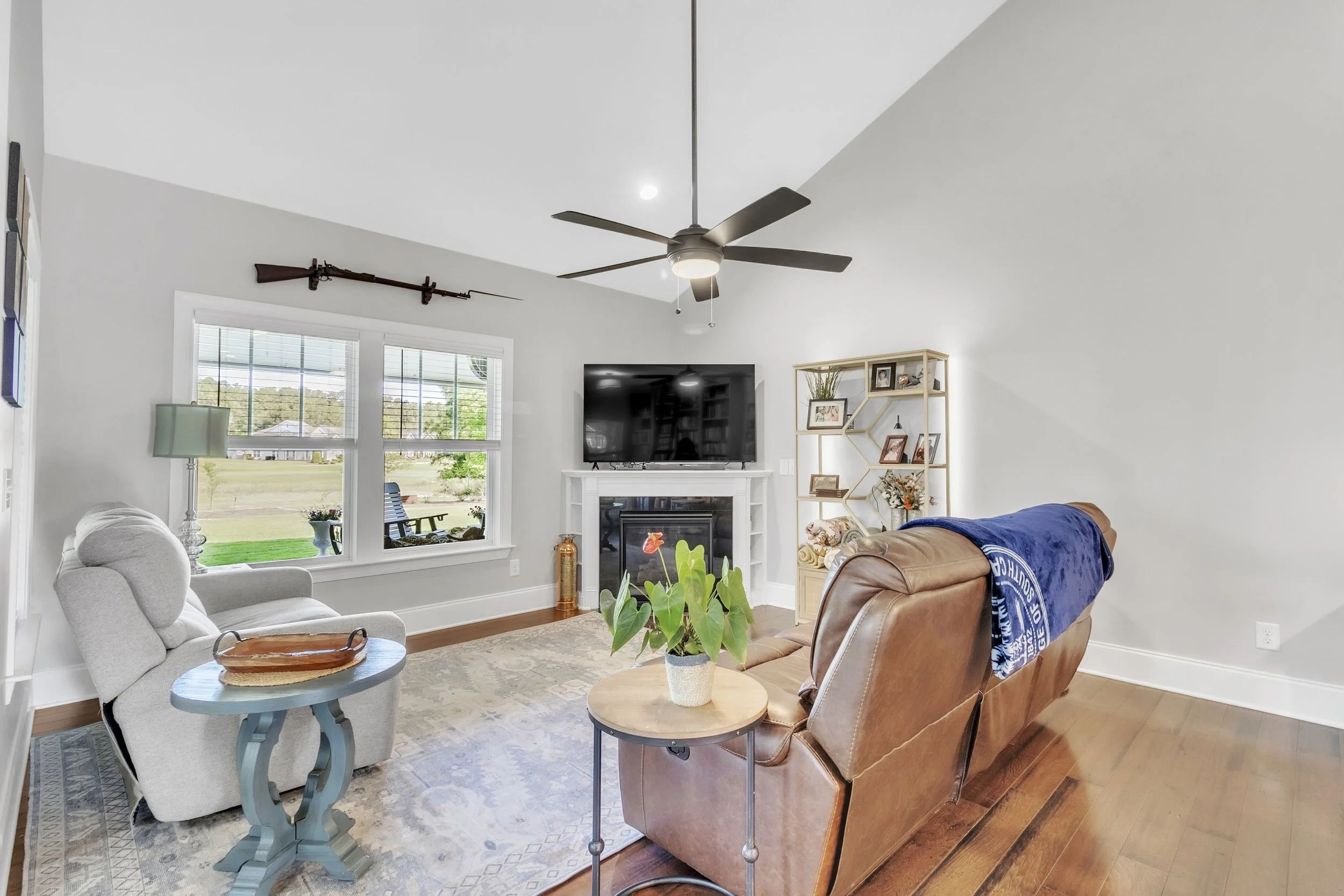 Living room with a ceiling fan, a large flat-screen TV above a fireplace, a tan leather sofa with a blue blanket, a white armchair, and a shelving unit with pictures and plants.