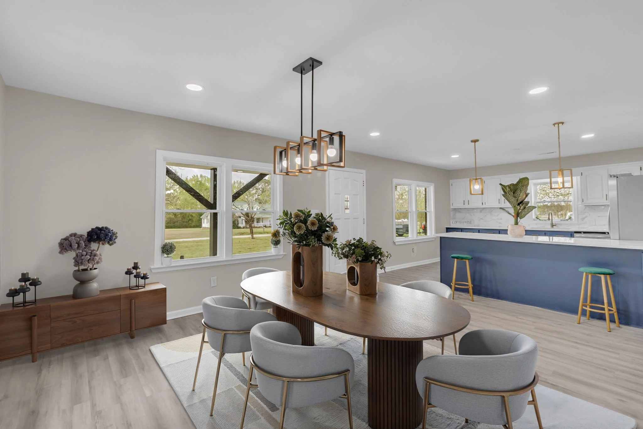 Open-concept living and kitchen area with dining table, gray chairs, wooden sideboard with floral arrangements, and kitchen bar with stools, large windows, and modern lighting fixtures.