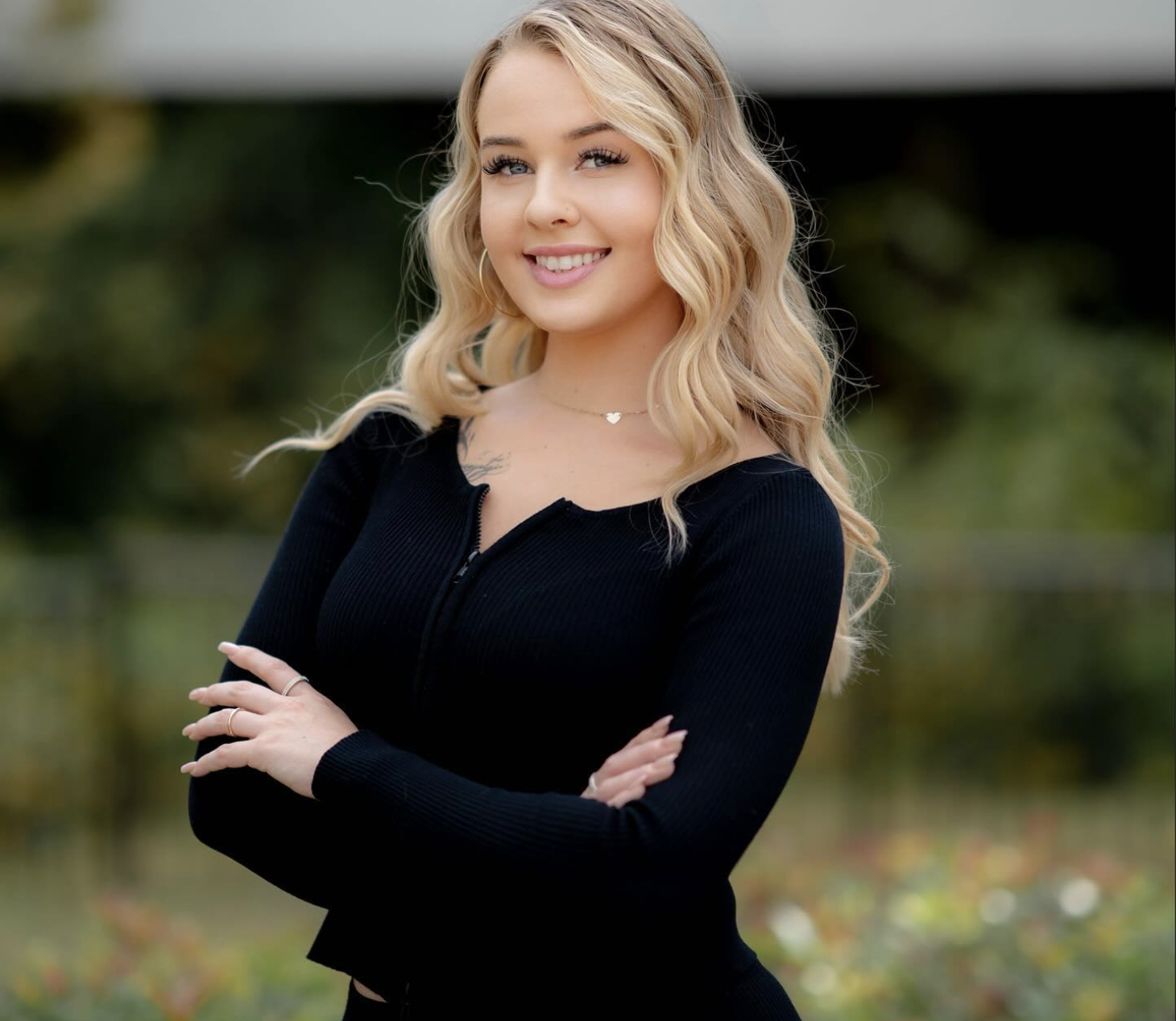 Young woman with blonde, wavy hair, wearing a black zip-up long sleeve top, outdoors with blurred nature background, smiling and crossing arms.