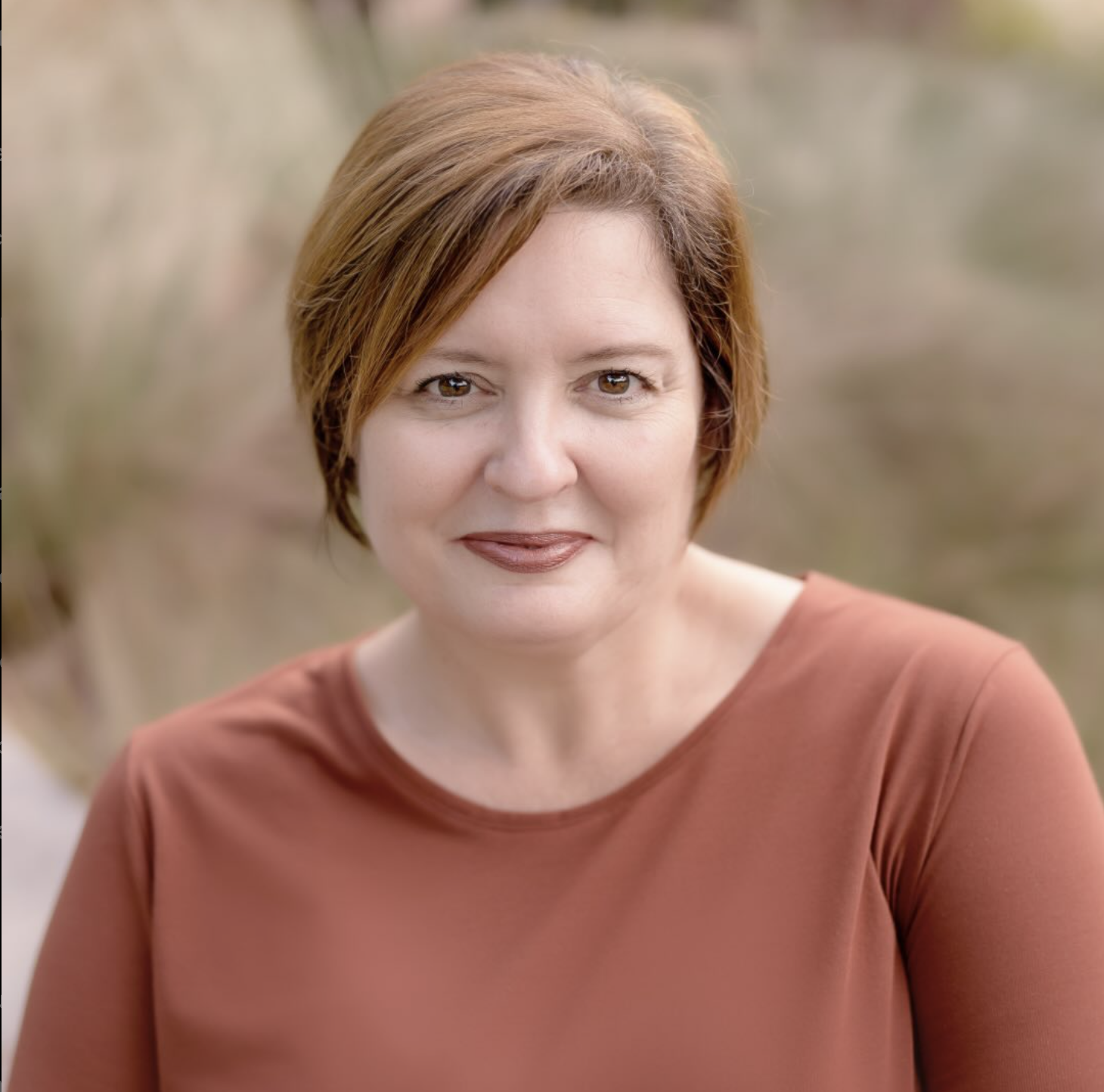 A woman with short red hair and brown eyes, wearing a rust-colored top, standing outdoors with a blurred natural background.