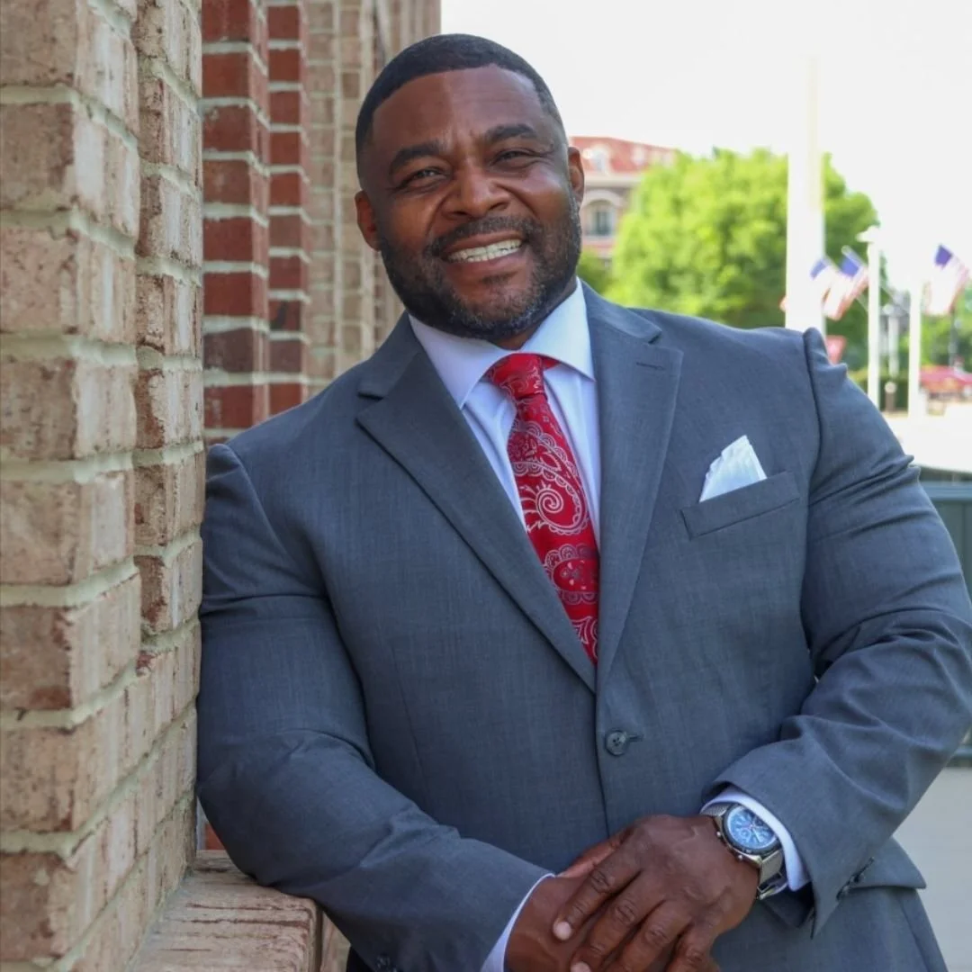 A woman with dark hair styled in loose curls wearing a bright orange blouse with ruffled collar, smiling, and wearing earrings, against a blurred background.