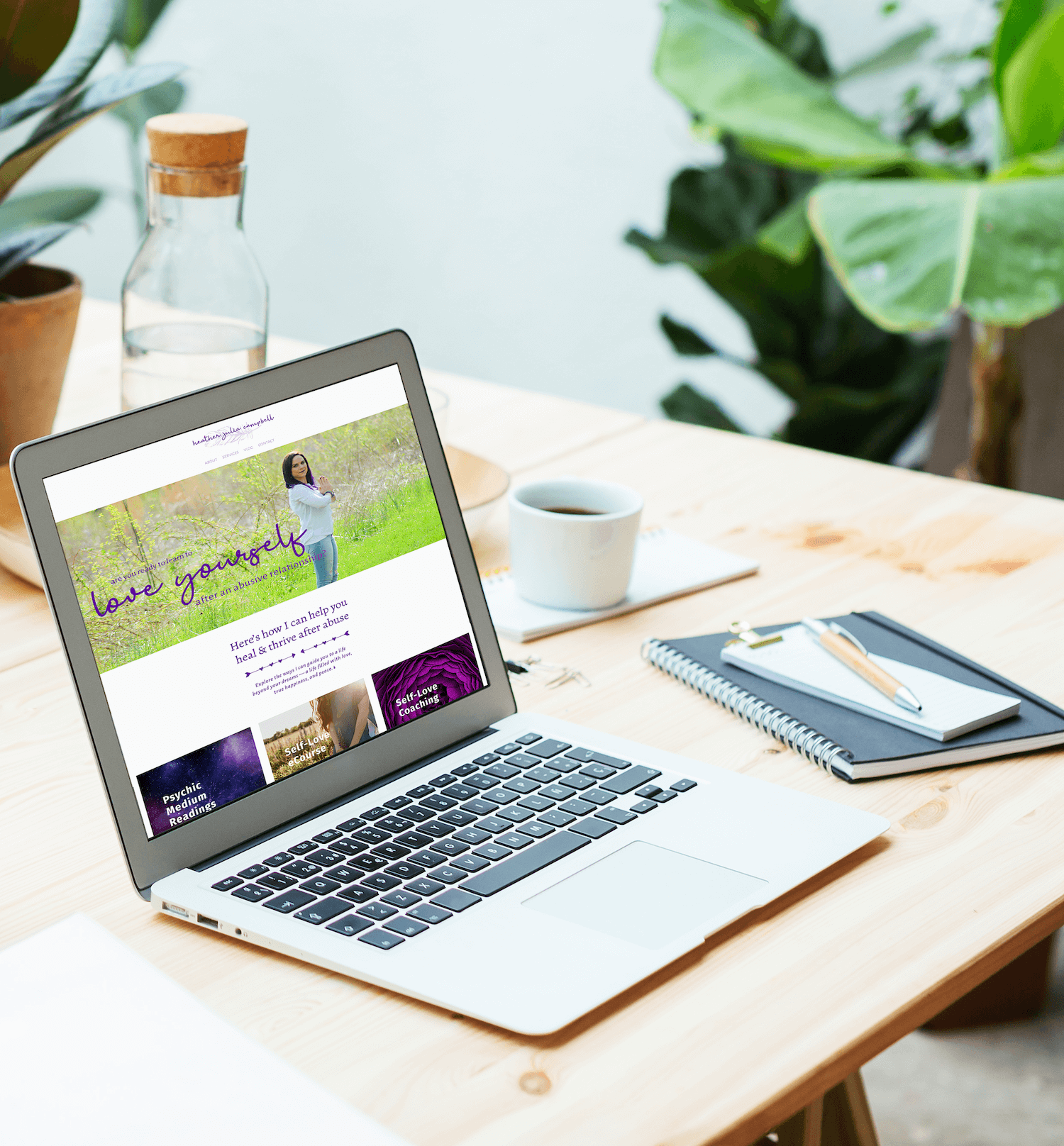 Laptop showing client website on table with notebook, mug, water jug and houseplant