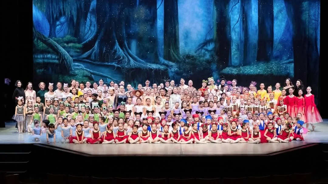 A large group of children and adults in colorful costumes on a theater stage with a forest background.