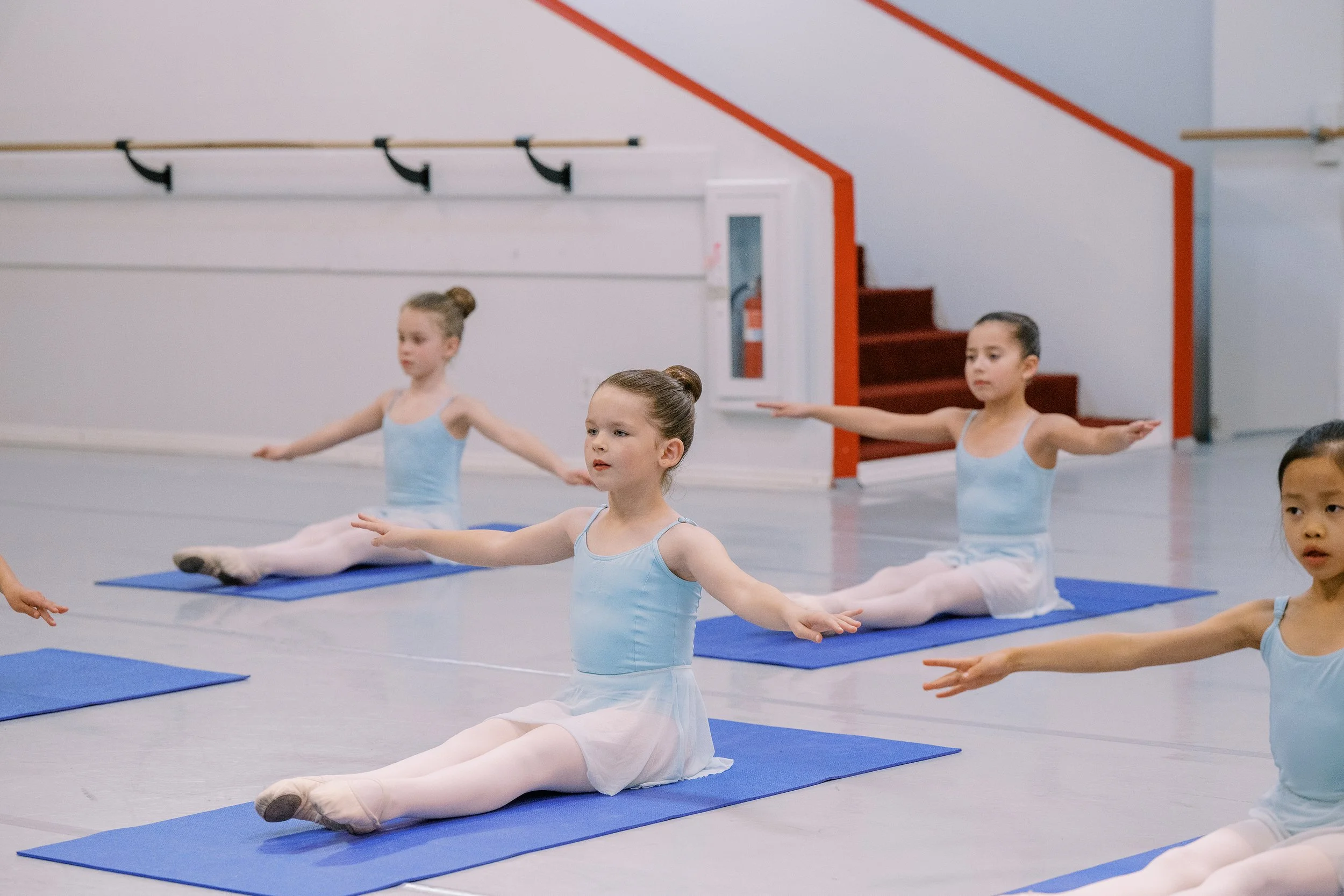 Young ballet students in a dance studio practicing seated stretches on blue mats.