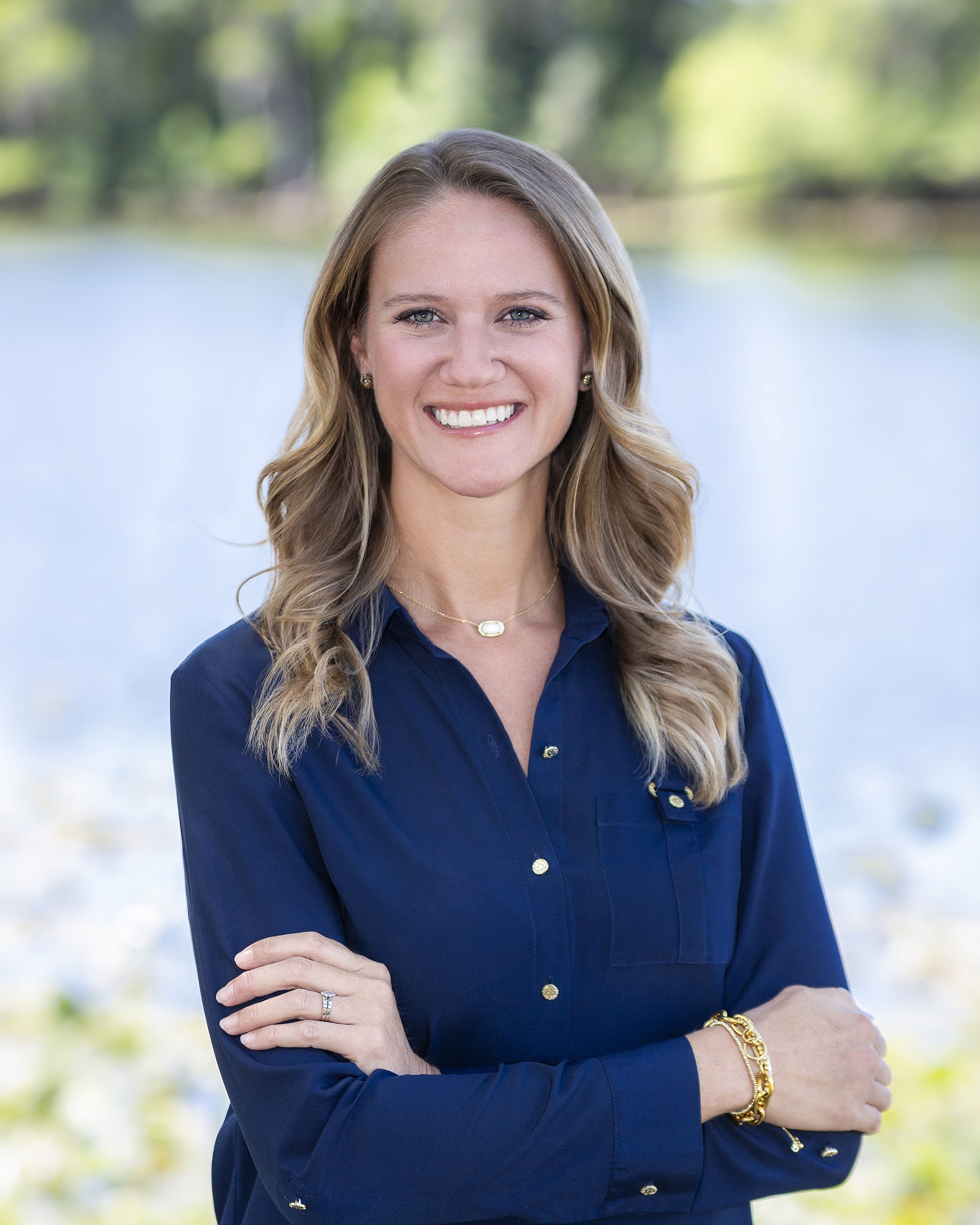 A smiling woman with long blonde hair wearing a navy blue shirt, gold jewelry, and standing outdoors with blurred green trees and water in the background.
