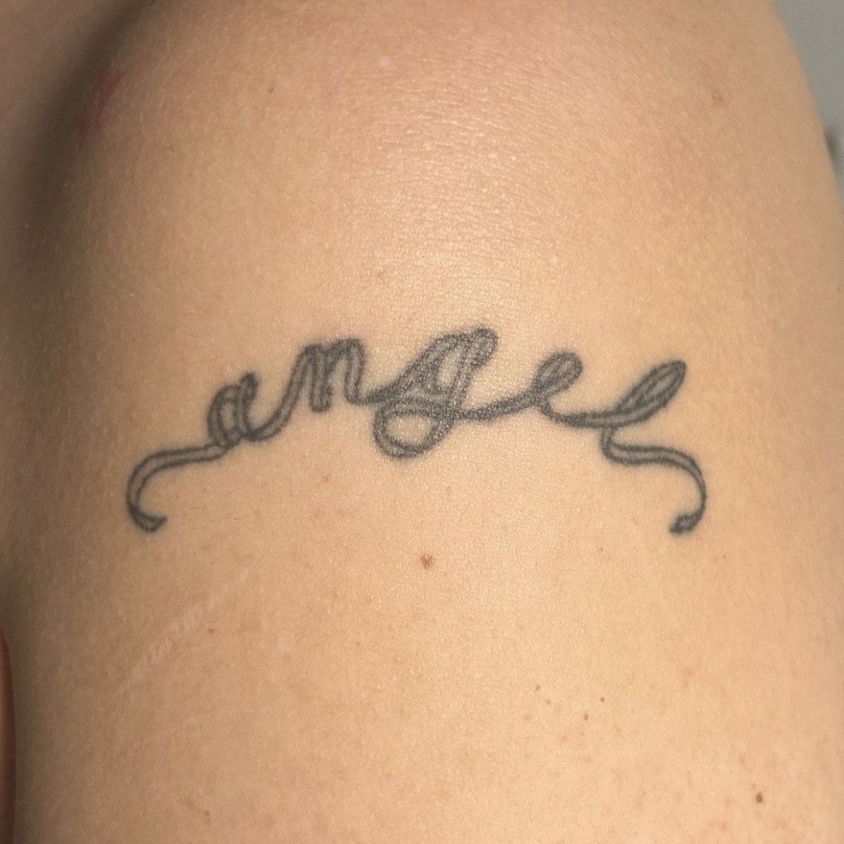 A tattoo with the word 'angel' written in cursive ribbon script on the arm