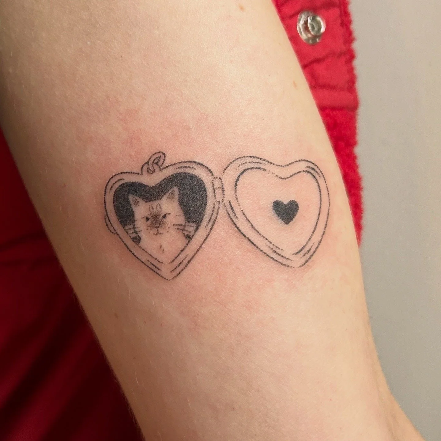 Tattoo of a heart locket featuring a cat portrait on one side and a small black heart on the other, on the arm