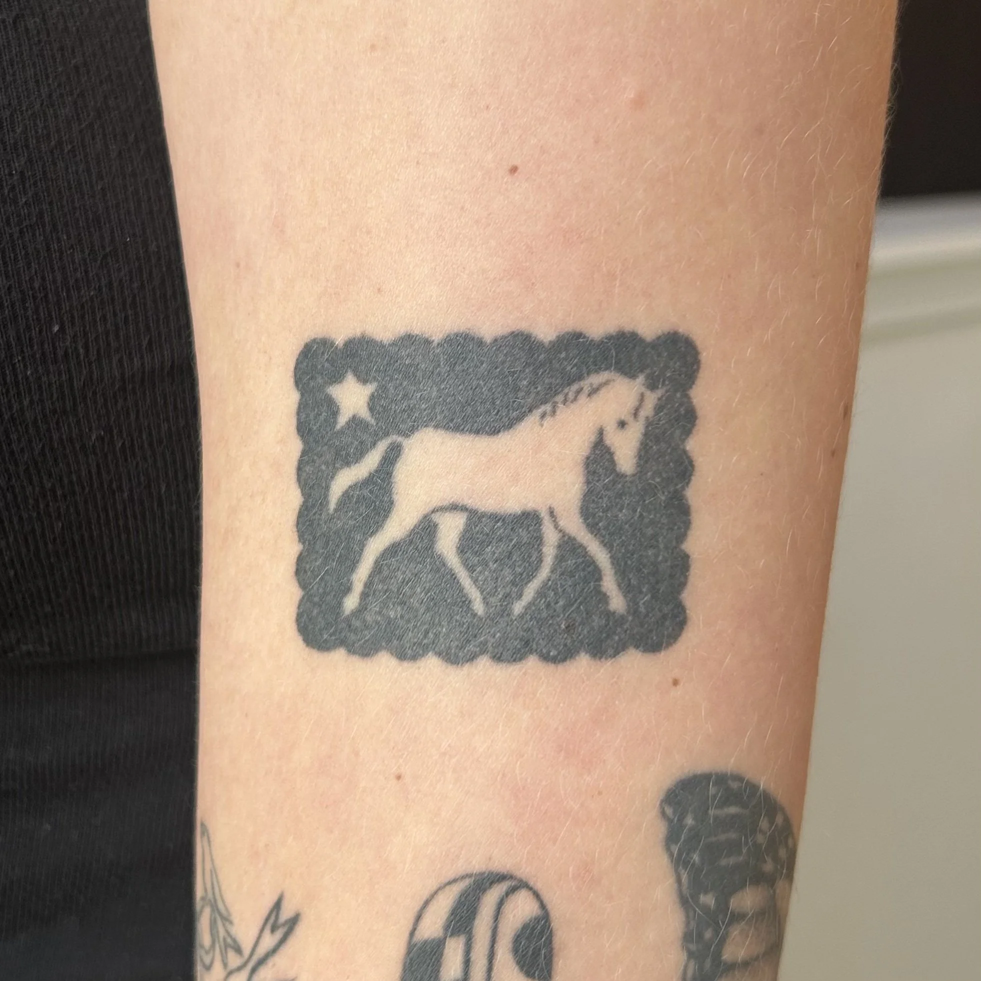 Tattoo of a white horse inside a black square with scalloped edges, with a star in the background on the arm