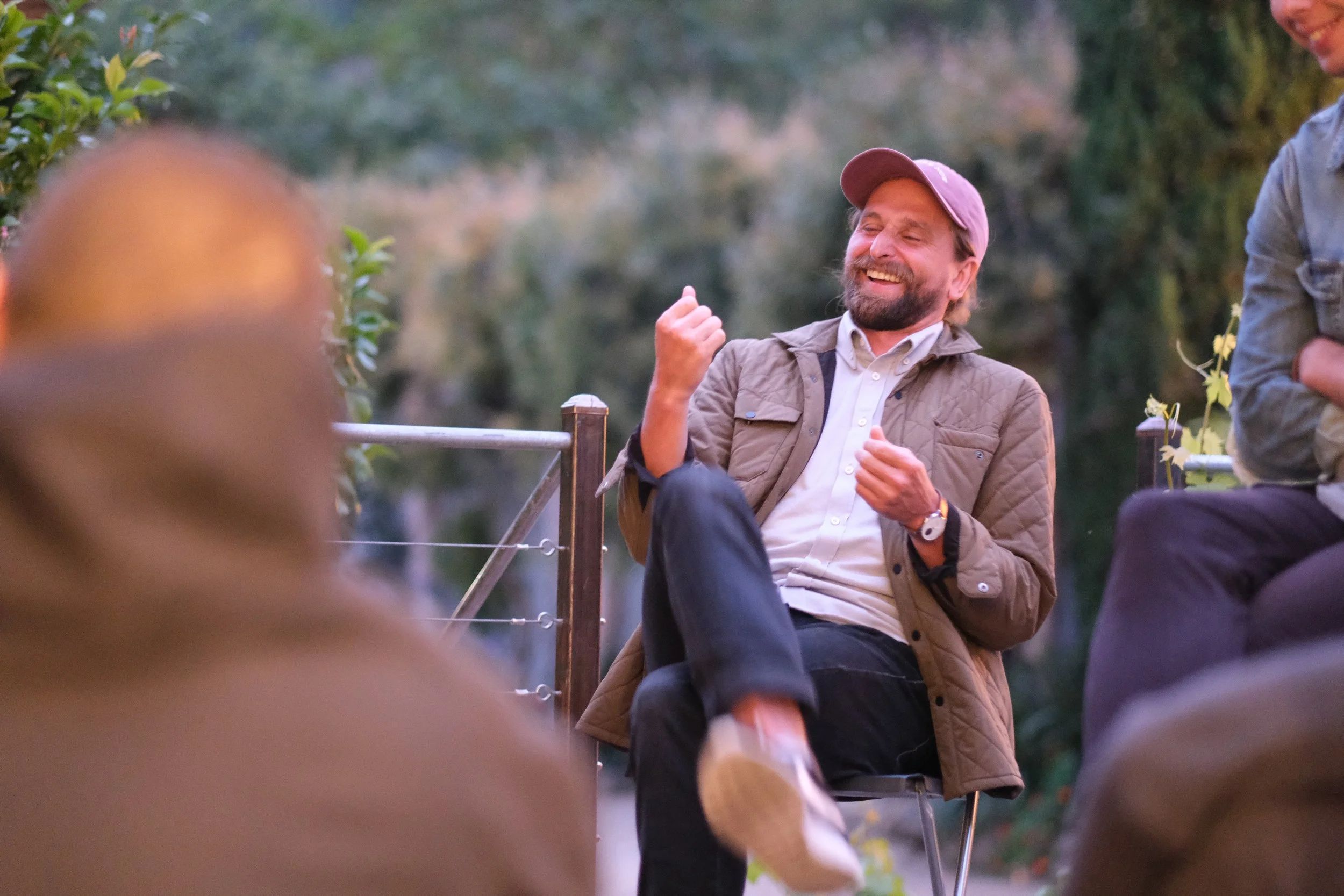 Man with beard in a pink cap and brown jacket smiling and gesturing during outdoor conversation with a group, with trees in the background.