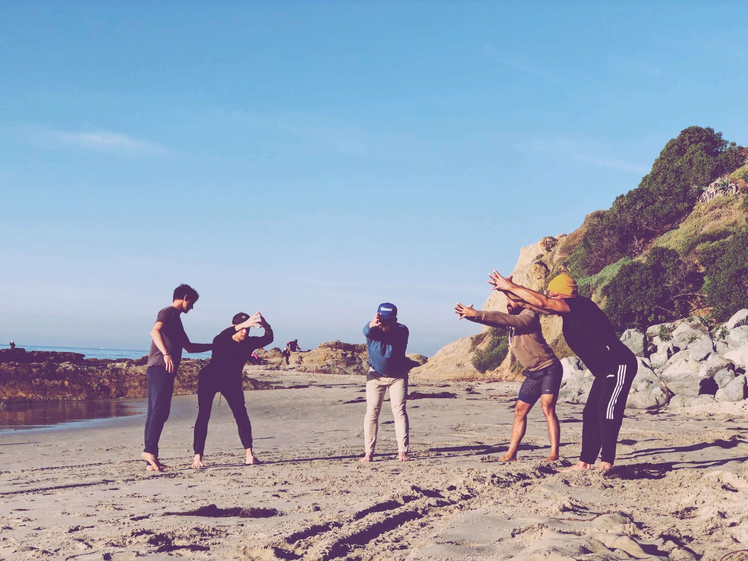 Group of friends at the beach taking photos, with cliffs and greenery in the background and clear blue sky.