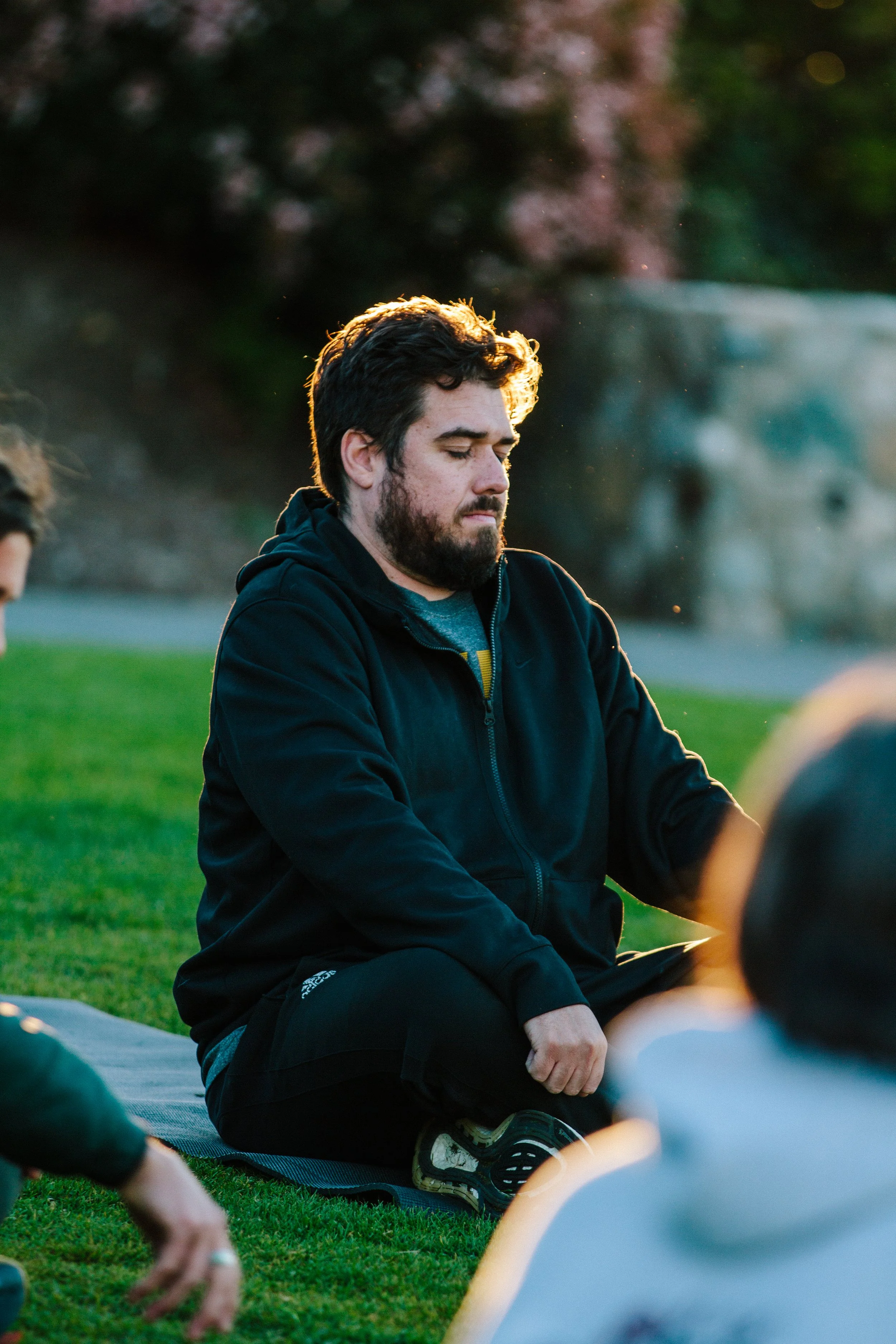 A man with dark hair and a beard sitting cross-legged on a yoga mat outdoors during sunset, with his eyes closed, in a meditative pose.