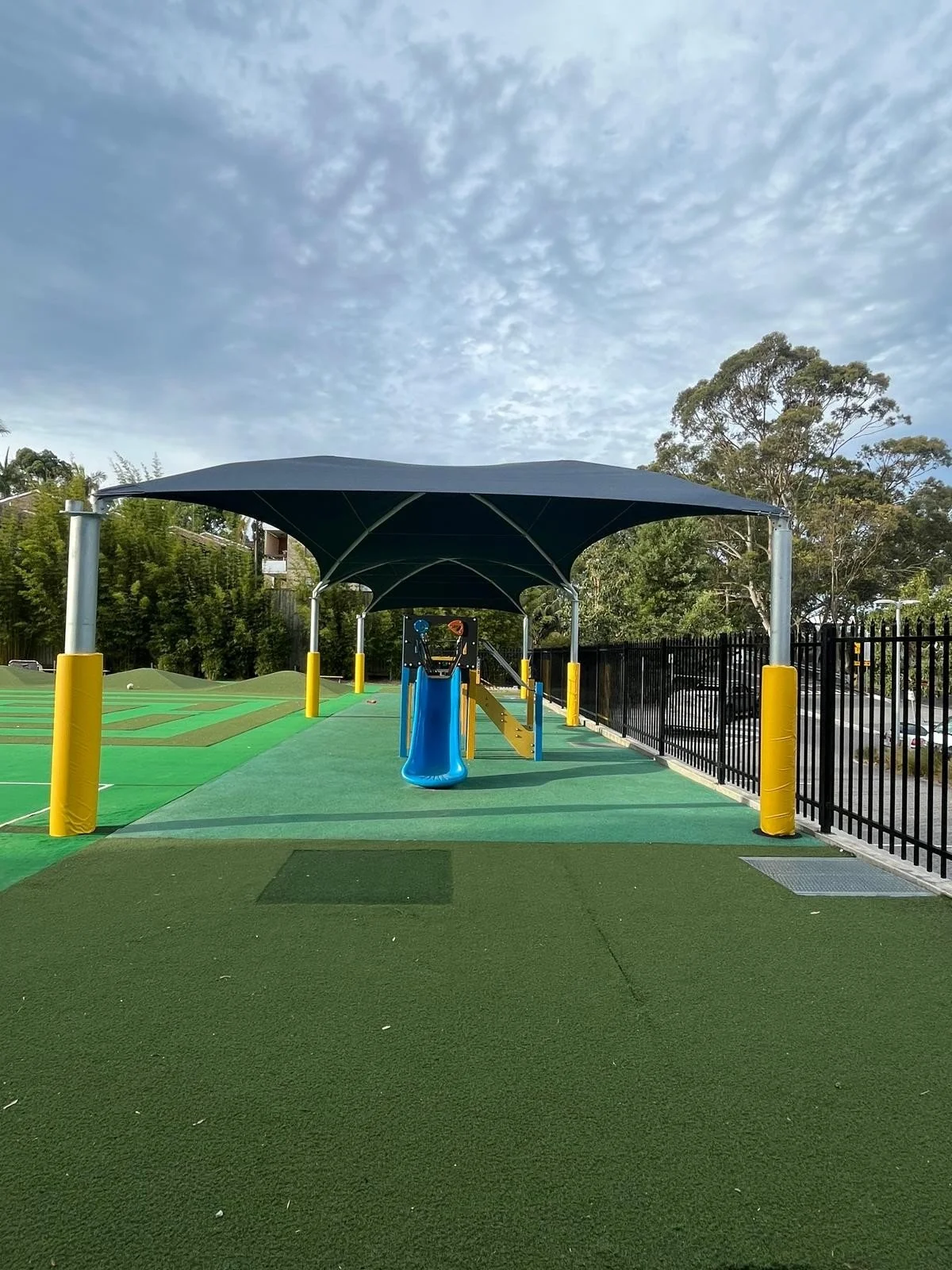 Playground with blue slide, shade cover, green and blue ground surfaces, black fence, and trees in the background under a partly cloudy sky.