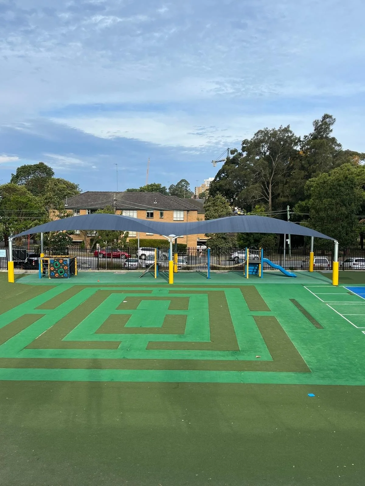 Children's playground area with a maze pattern on the ground, covered by a large shade structure. There are play structures including slides and climbing equipment, surrounded by a metal fence, with trees and houses in the background.