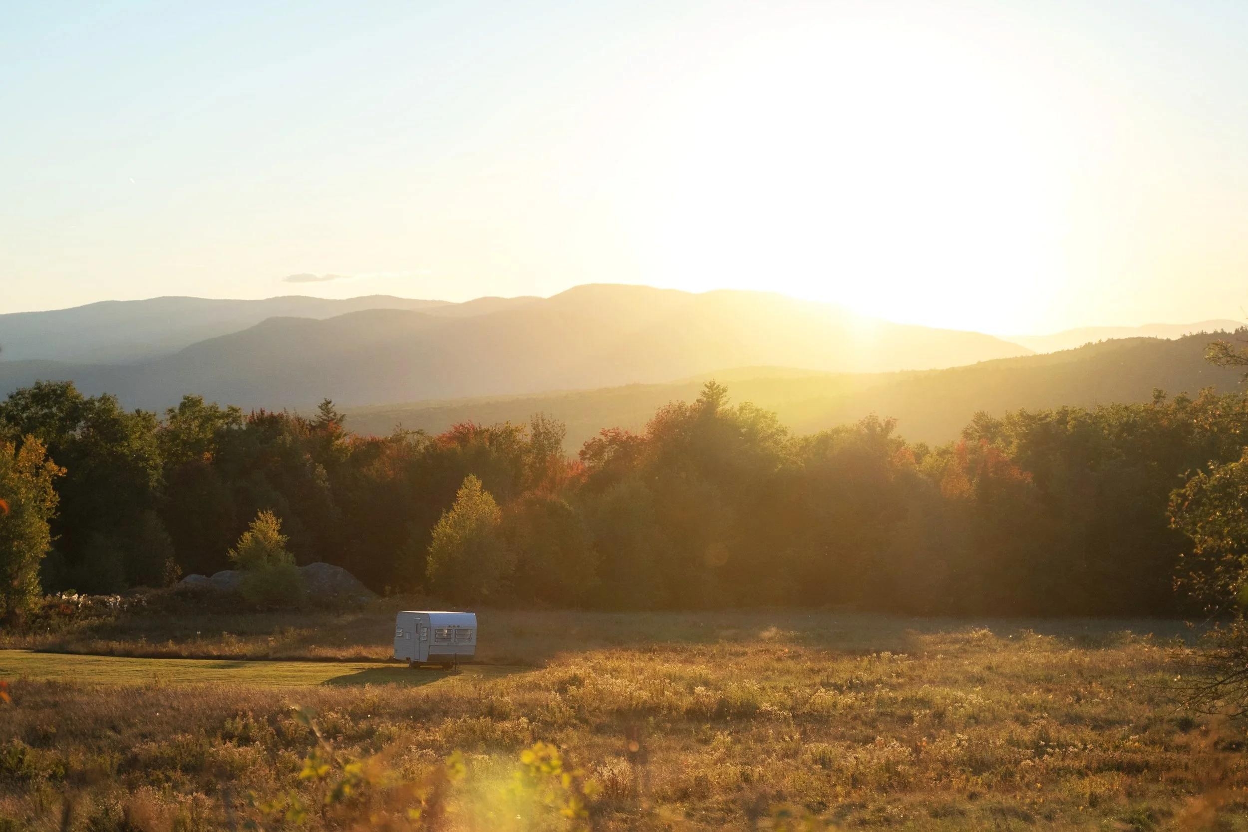 Sunset over a landscape with mountains, trees, and a small camper trailer in an open field.