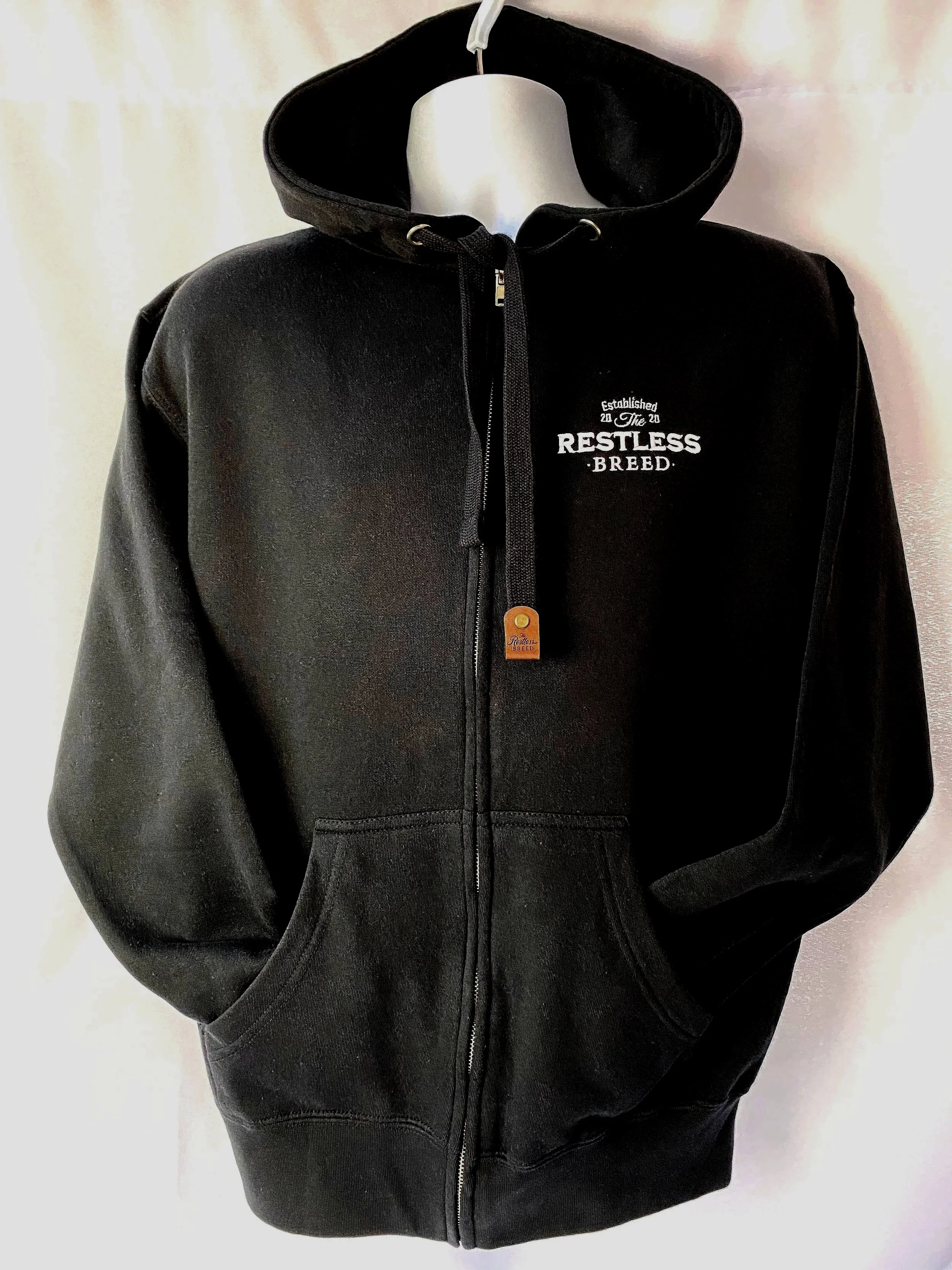 The restless breed hooded zip up black sweatshirt with left pocket embroidered logo and signature leatherette tab on drawstring