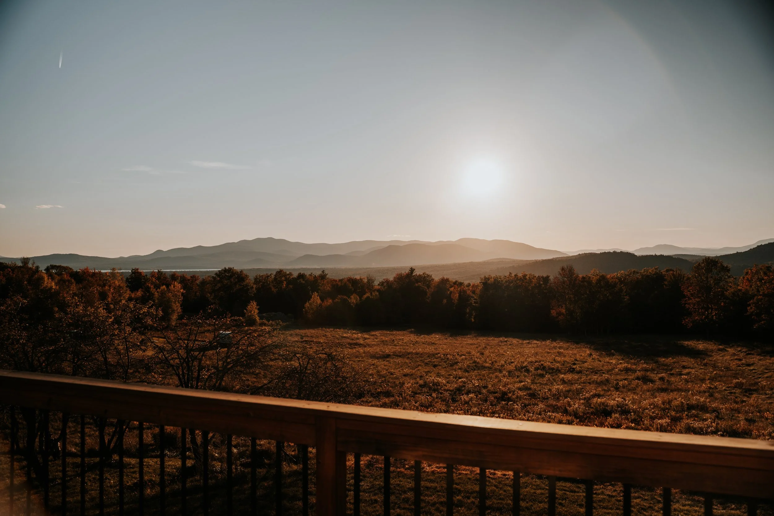 Scenic view of mountains, trees, and fields at sunset, taken from a balcony with railing.