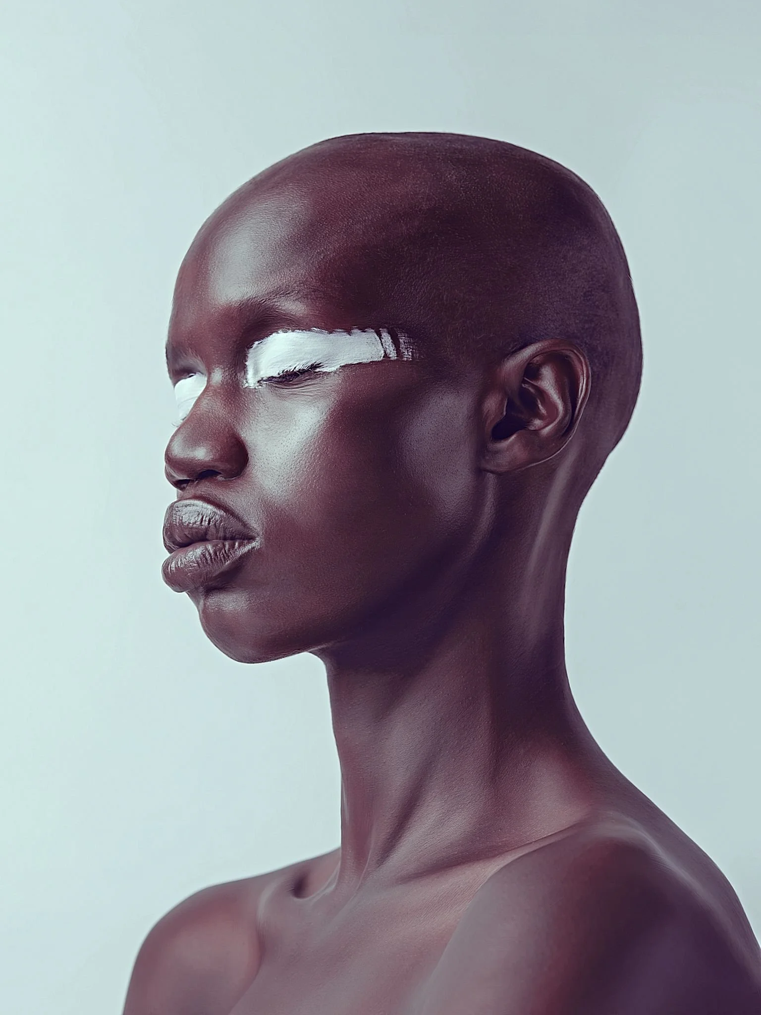 A close-up portrait of a dark-skinned woman with a shaved head and white artistic makeup across her eyes.