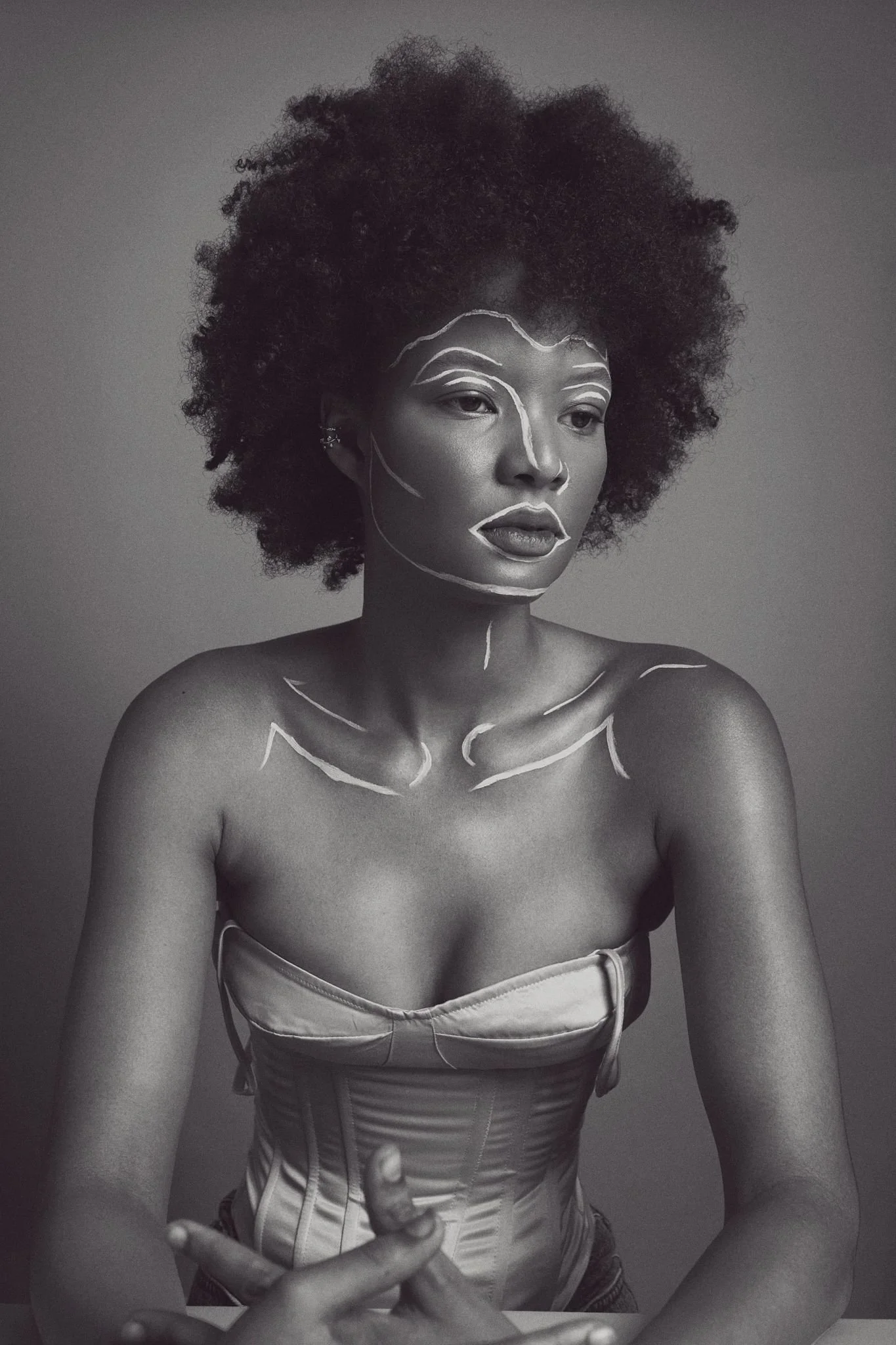 Black and white portrait of a woman with natural curly hair, wearing a strapless top and a corset, with white lines drawn on her face and shoulders, against a plain gray background.