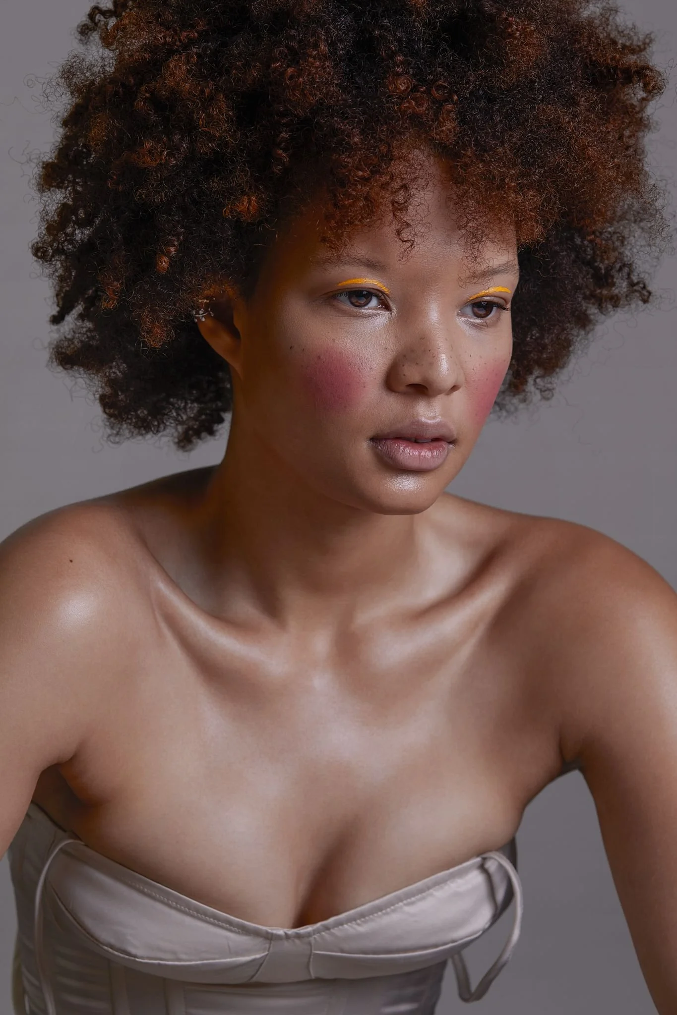 A woman with curly, reddish-brown hair looking down slightly against a plain gray background. She has pink blush, yellow eye makeup, and wears a strapless, beige-colored top.