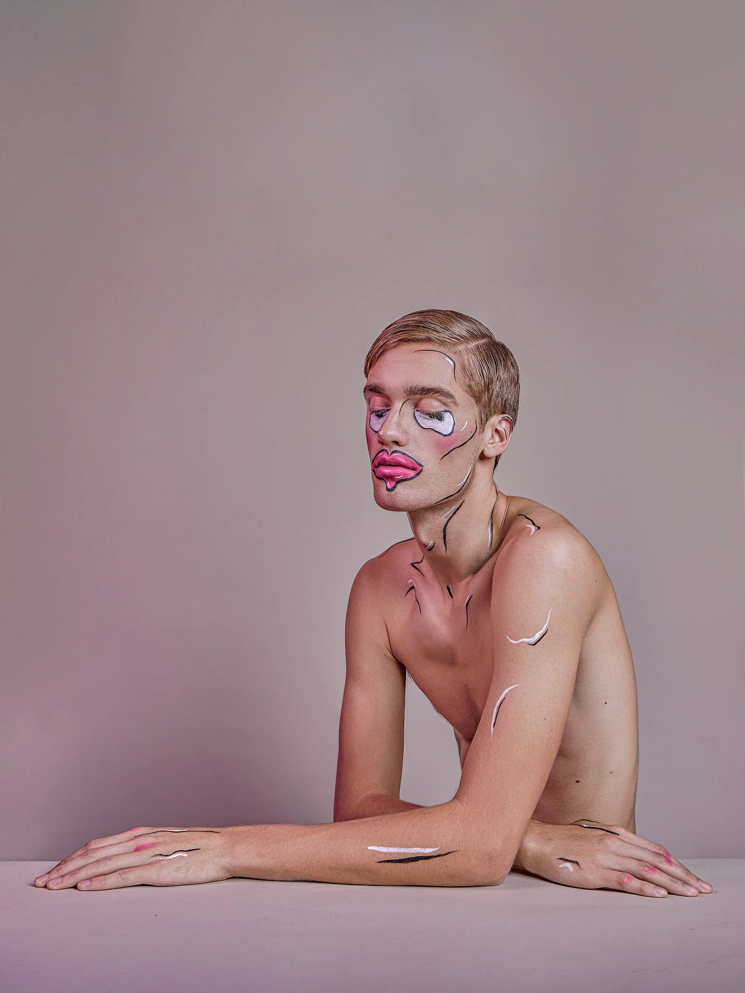 A shirtless person with short hair is sitting at a table with their arms crossed, with colorful paint and comic-style outlines on their face and body, against a plain background.