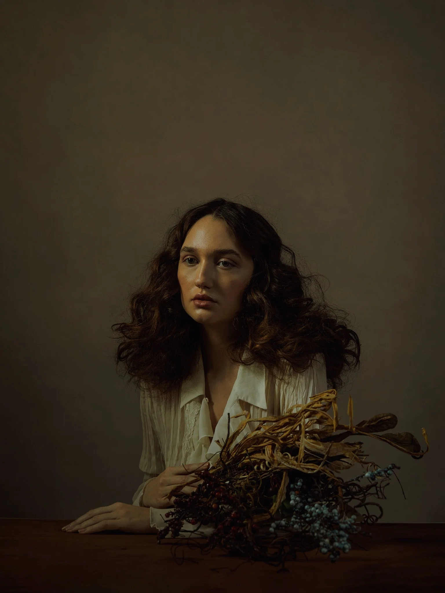 A woman with curly hair and a neutral expression, wearing a white blouse, sitting at a table with a dried flower arrangement in front of her, against a plain, dark background.