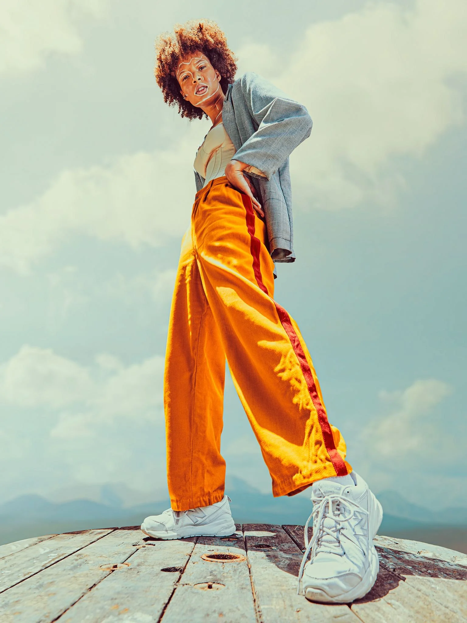 A woman with curly hair wearing a gray blazer, beige top, bright orange wide-leg pants with red stripe, and white sneakers standing on a wooden platform outdoors.