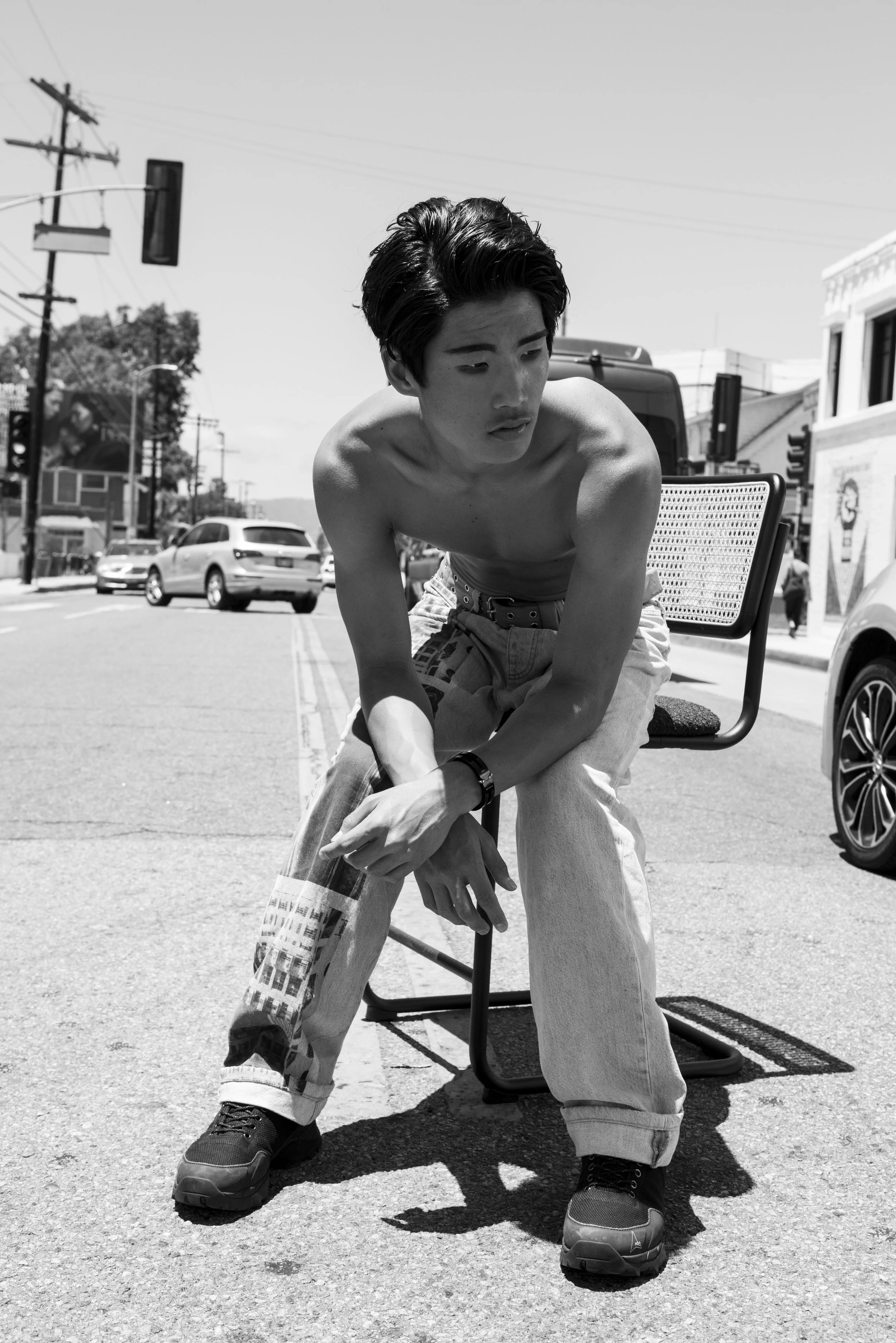 A shirtless young man with dark hair sitting on a folding chair on a city street, looking down, with cars and buildings in the background.