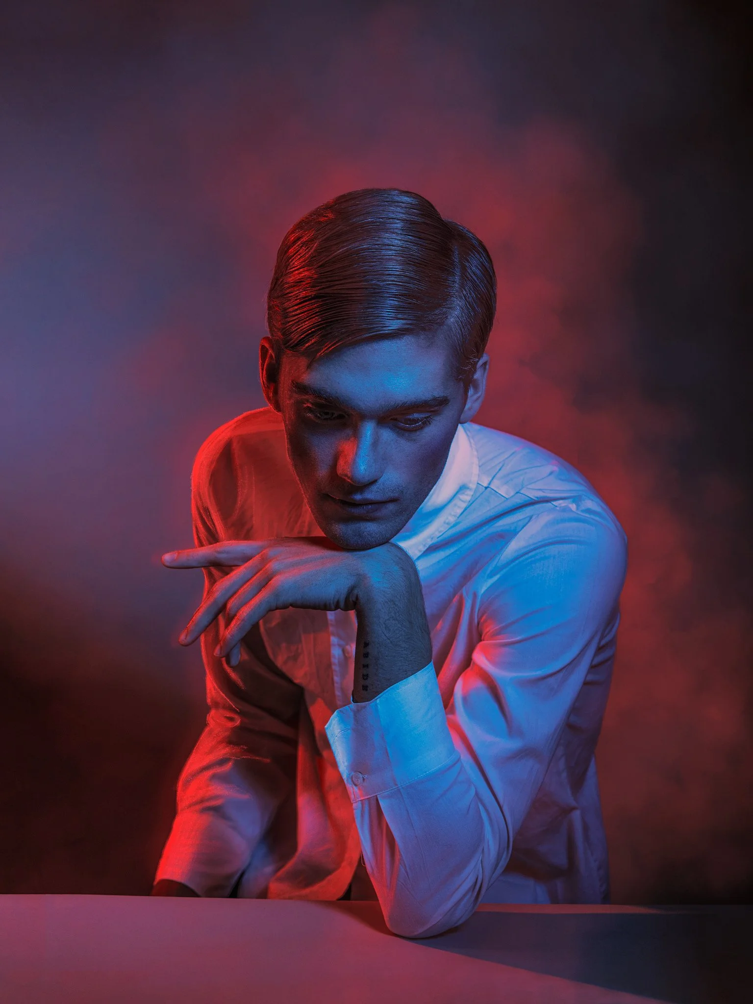 A young man with slicked-back hair wearing a white shirt, resting his chin on his hand, with a serious expression against a dark background with red and blue lighting.