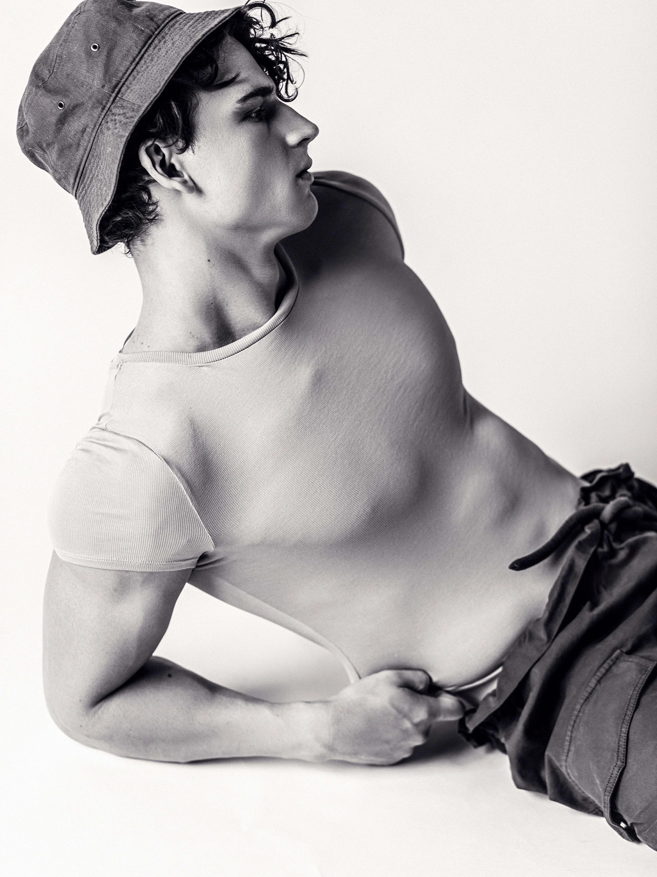 Black and white photo of a young man with curly hair wearing a bucket hat, sitting with one arm supporting his body, wearing a fitted T-shirt and drawstring shorts.