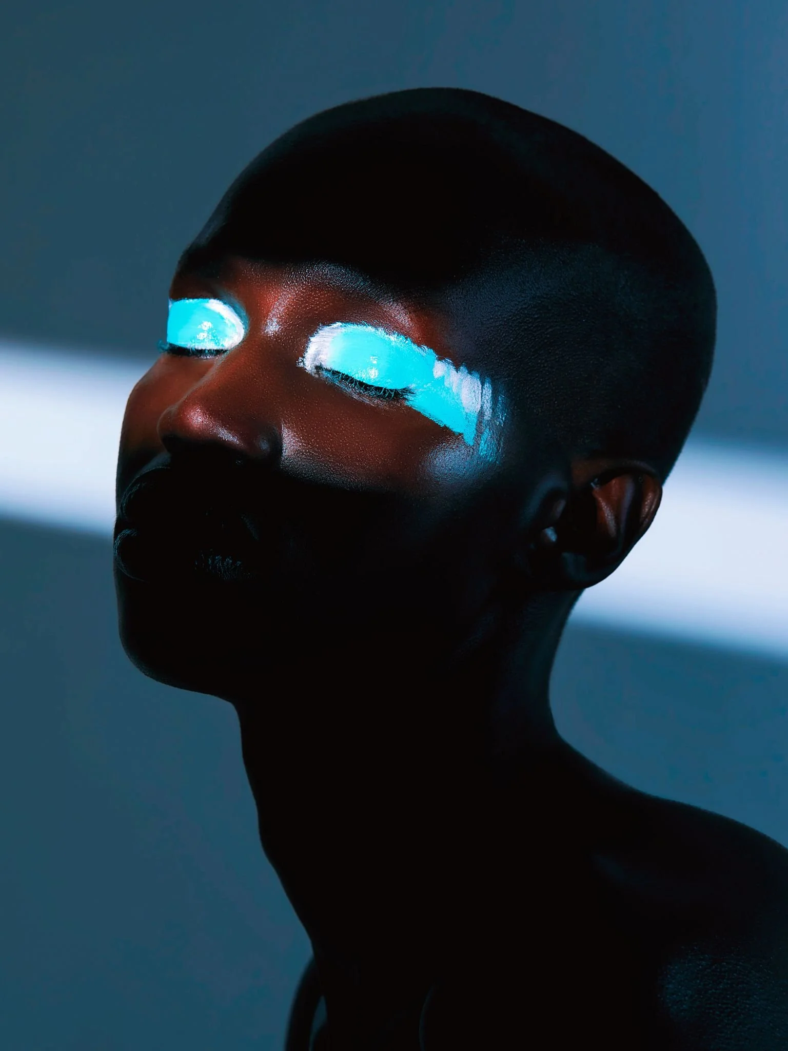 A person with short hair and dark skin has bright blue eye makeup that extends across the eyelids and slightly beyond, against a dark background.