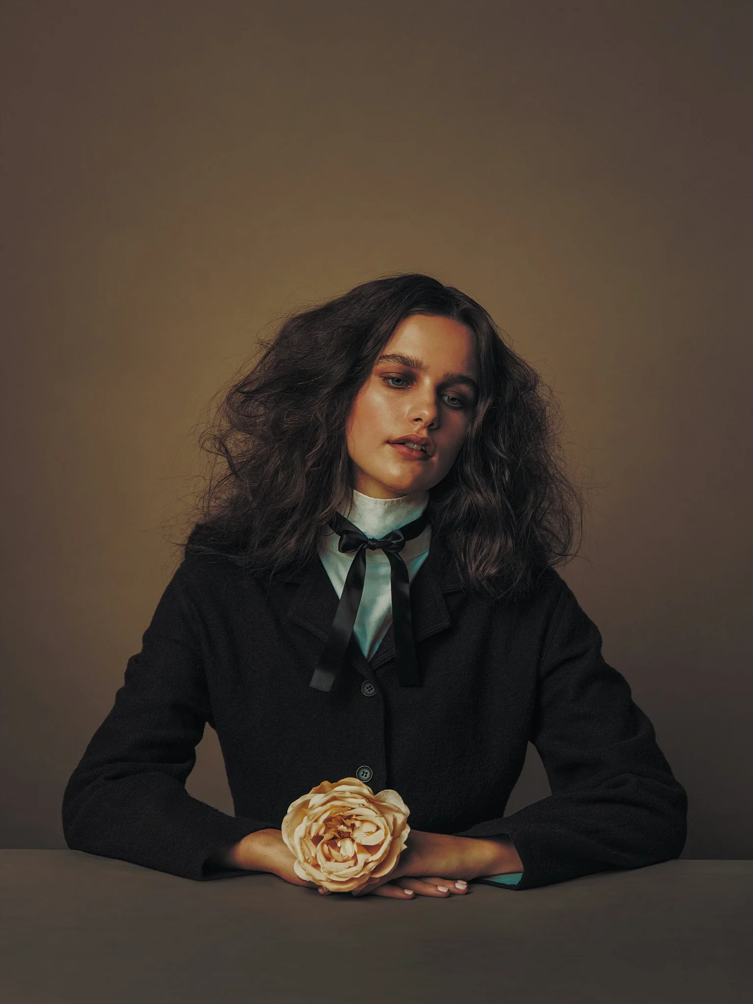 Portrait of a woman with dark curly hair, wearing a black jacket, white shirt with a black ribbon, sitting at a table with a large beige flower in front of her against a plain, muted background.