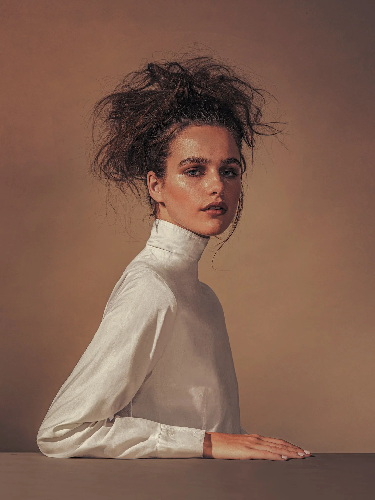 A woman with curly, messy hair wearing a high-neck white satin blouse, sitting with her arm resting on a surface against a plain, warm-toned background.