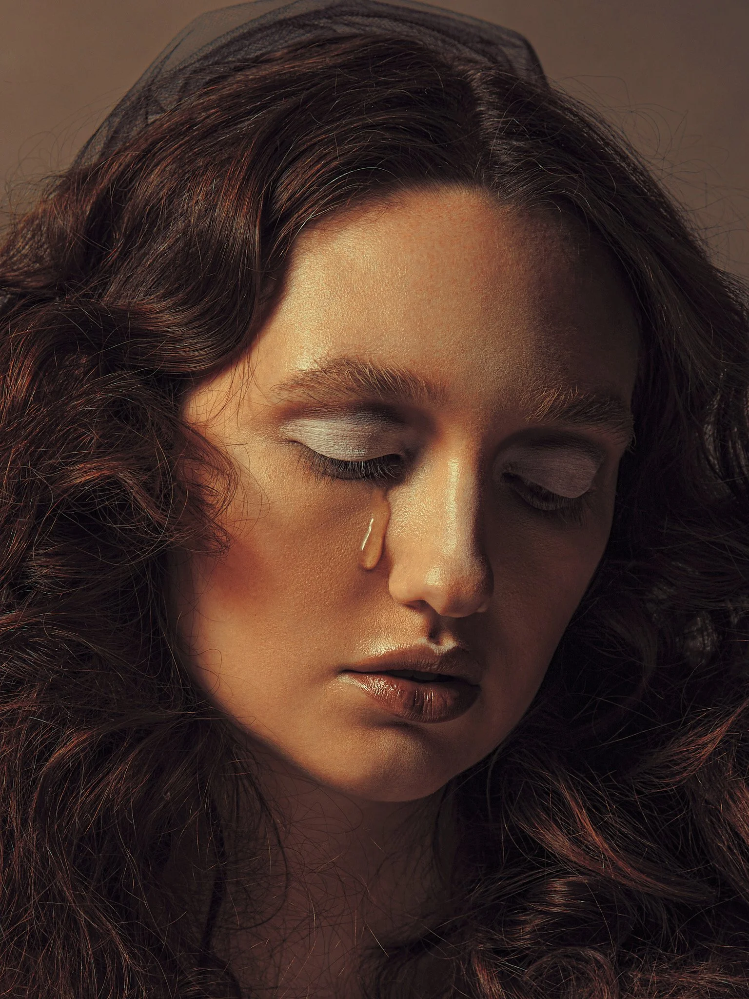 Close-up portrait of a woman with long, wavy brown hair, light makeup, and her eyes closed, showing a tear rolling down her cheek.