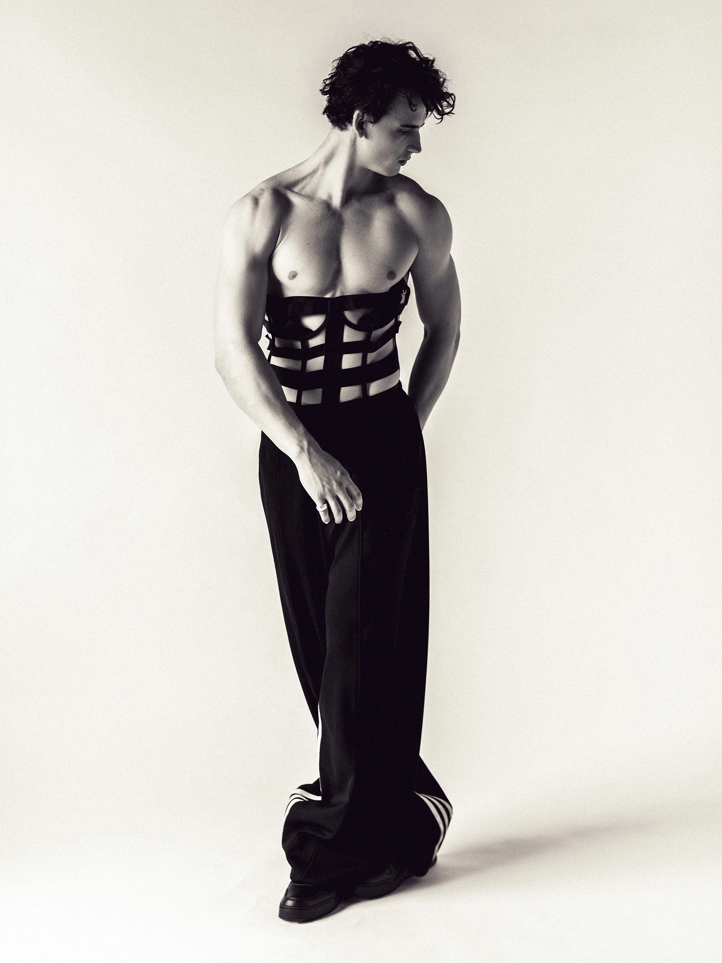 A man with messy hair is standing against a plain light background, wearing black pants and a harness-style top that exposes his chest."