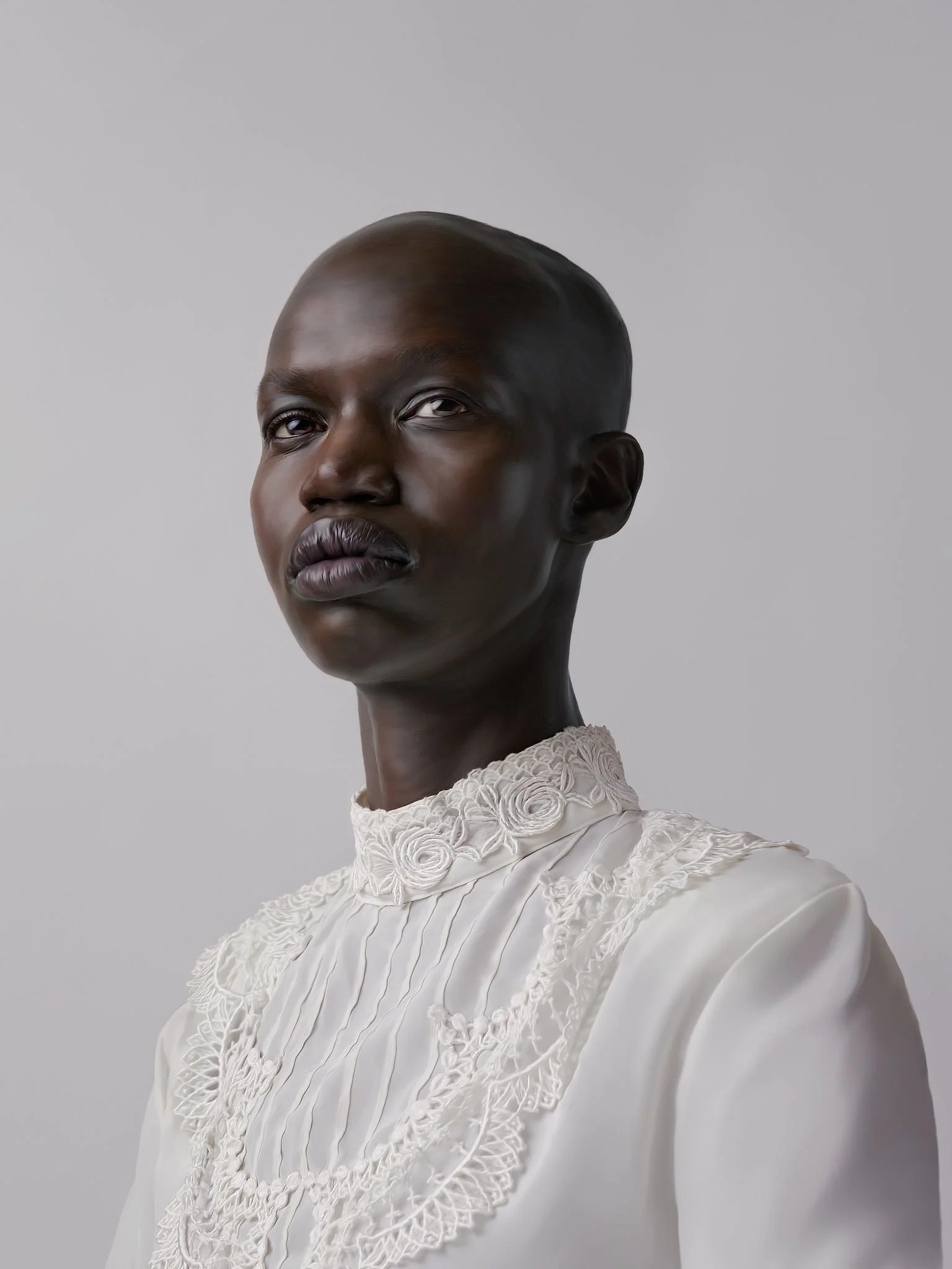 Portrait of a dark-skinned woman with a shaved head, wearing a white embroidered top against a plain gray background.