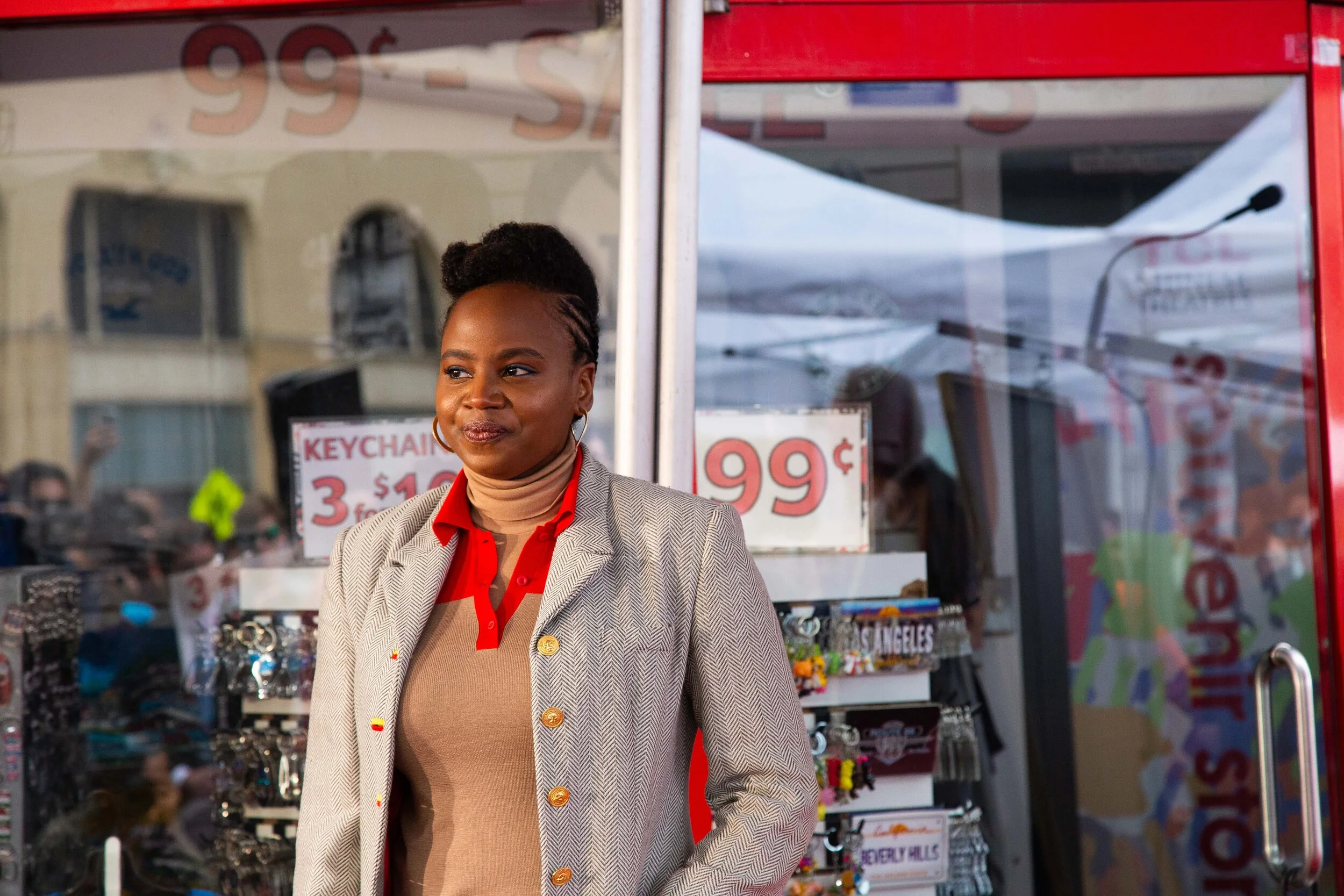 Director Dee Rees standing in front of a store window with sale signs. She has braided hair, wearing a beige blazer with gold buttons, a red shirt underneath, and a beige turtleneck. She appears to be posing or waiting outside a retail shop.