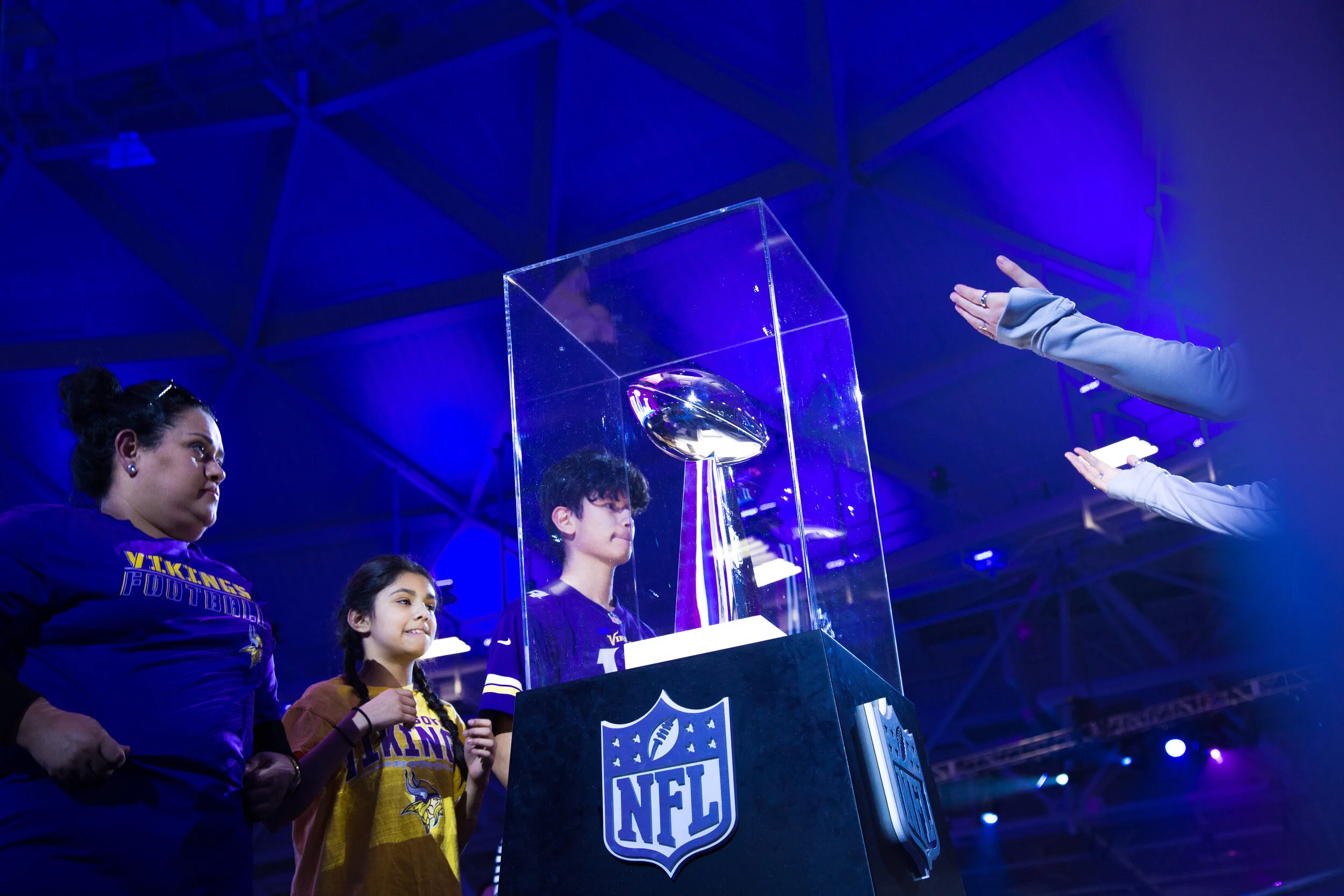 Young football fans, including two women and a girl, observe the Vince Lombardi Trophy displayed in a glass case at an NFL Superbowl LII event, with a blue and purple lighting in the background.