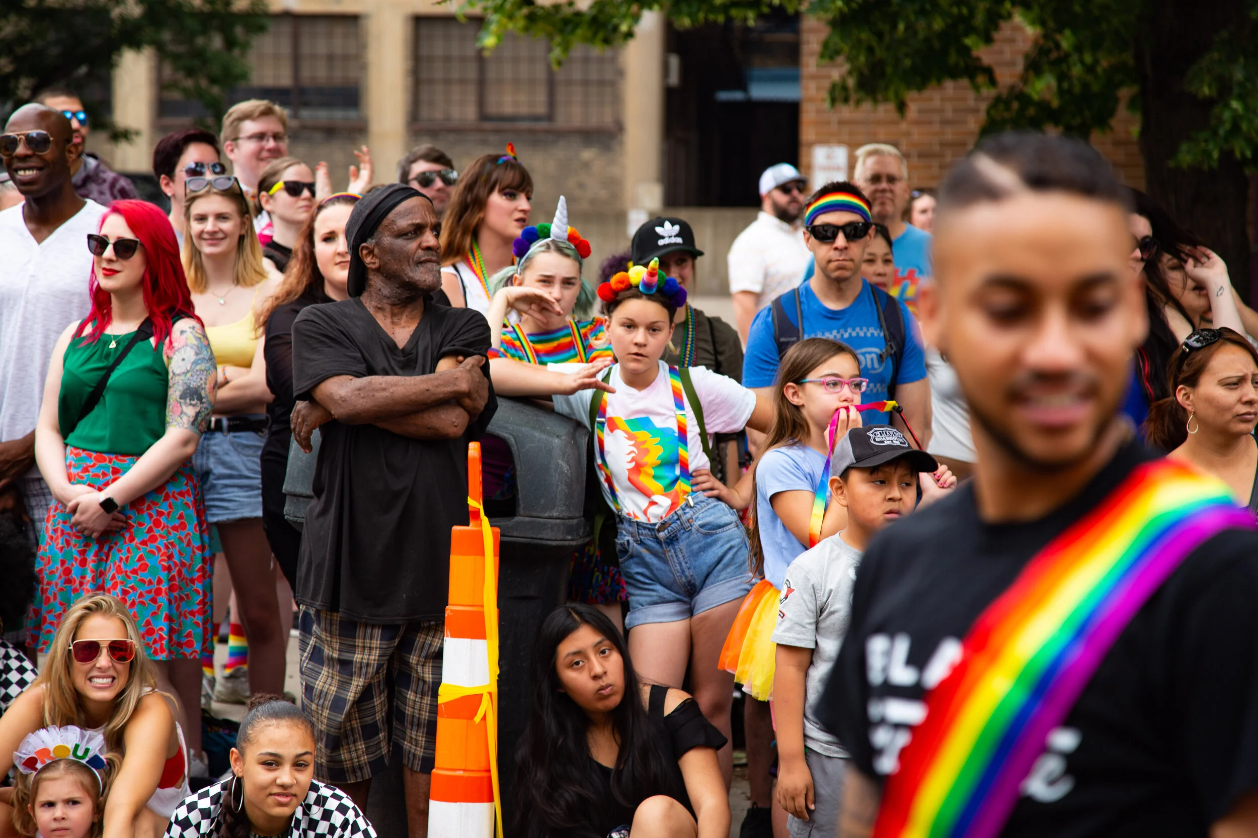 A diverse group of people gathered outdoors during a pride parade, many wearing rainbow accessories and colorful clothing, with some children sitting on the ground and adults standing in the background.