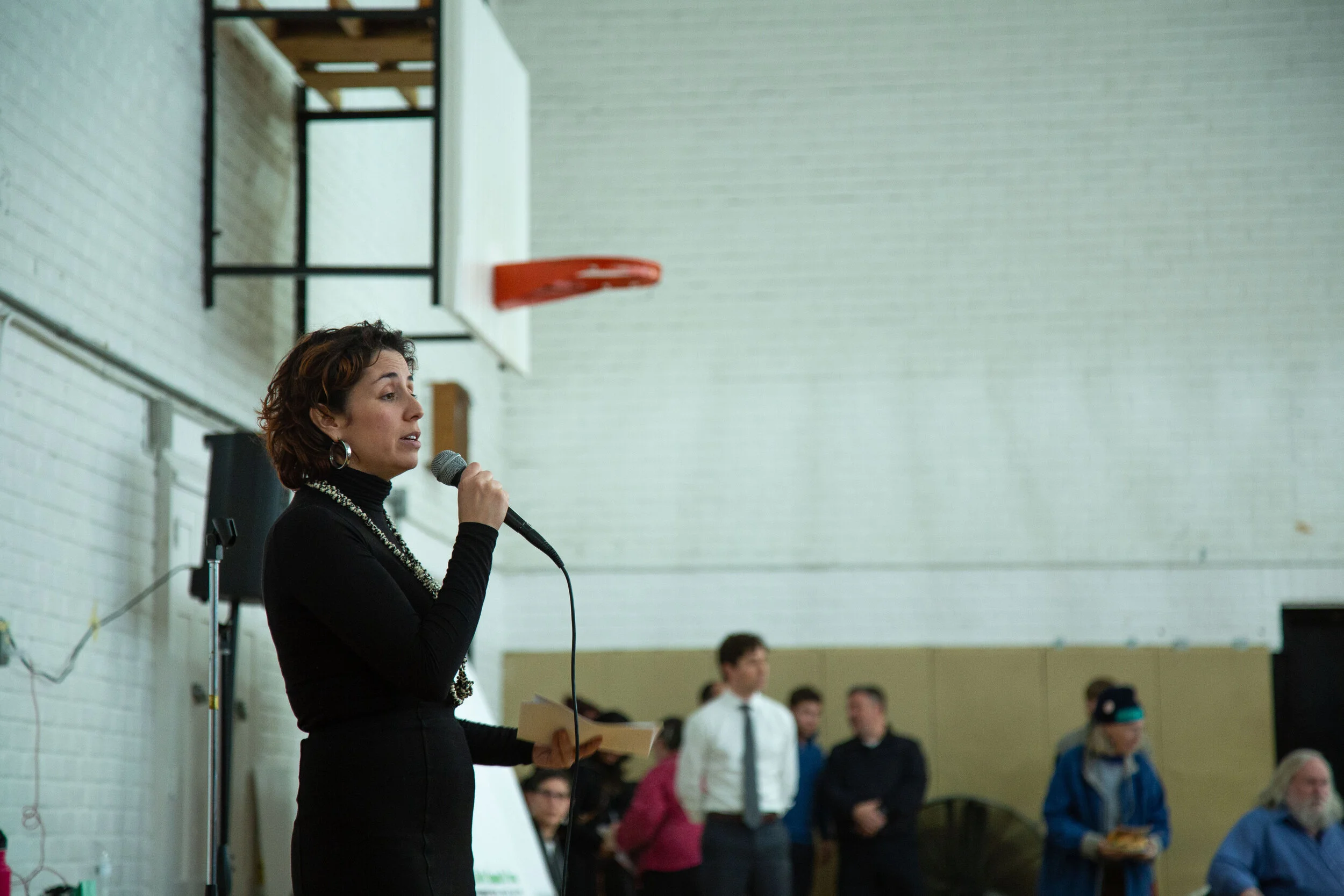 A woman with short dark hair, wearing a black turtleneck and jewelry, speaking into a microphone in a gymnasium, with a group of people standing behind her.