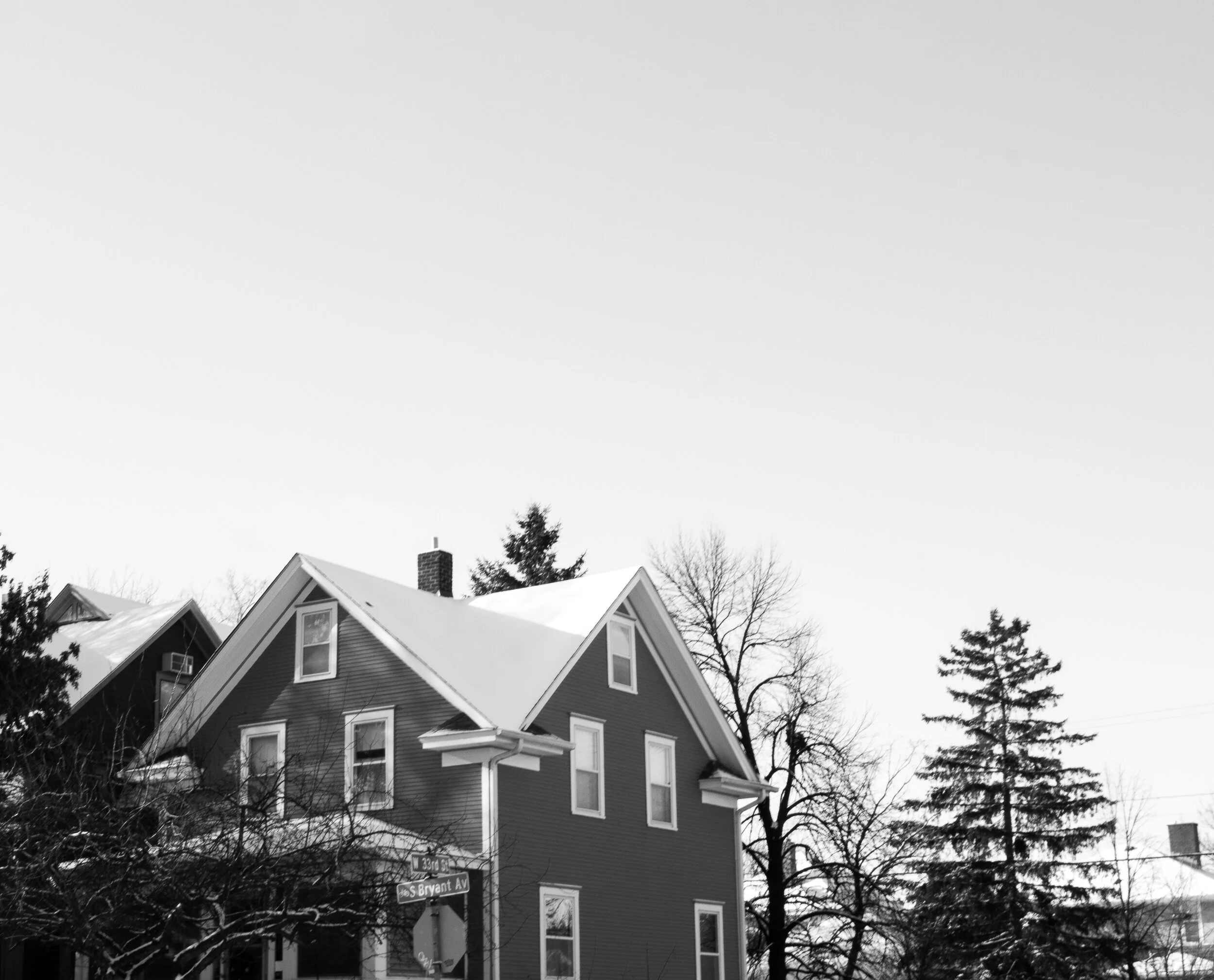 A black and white photo of a house with a steep roof and multiple windows, trees without leaves, and a street sign indicating '135th St' and 'Bryant Ave' in a winter setting.