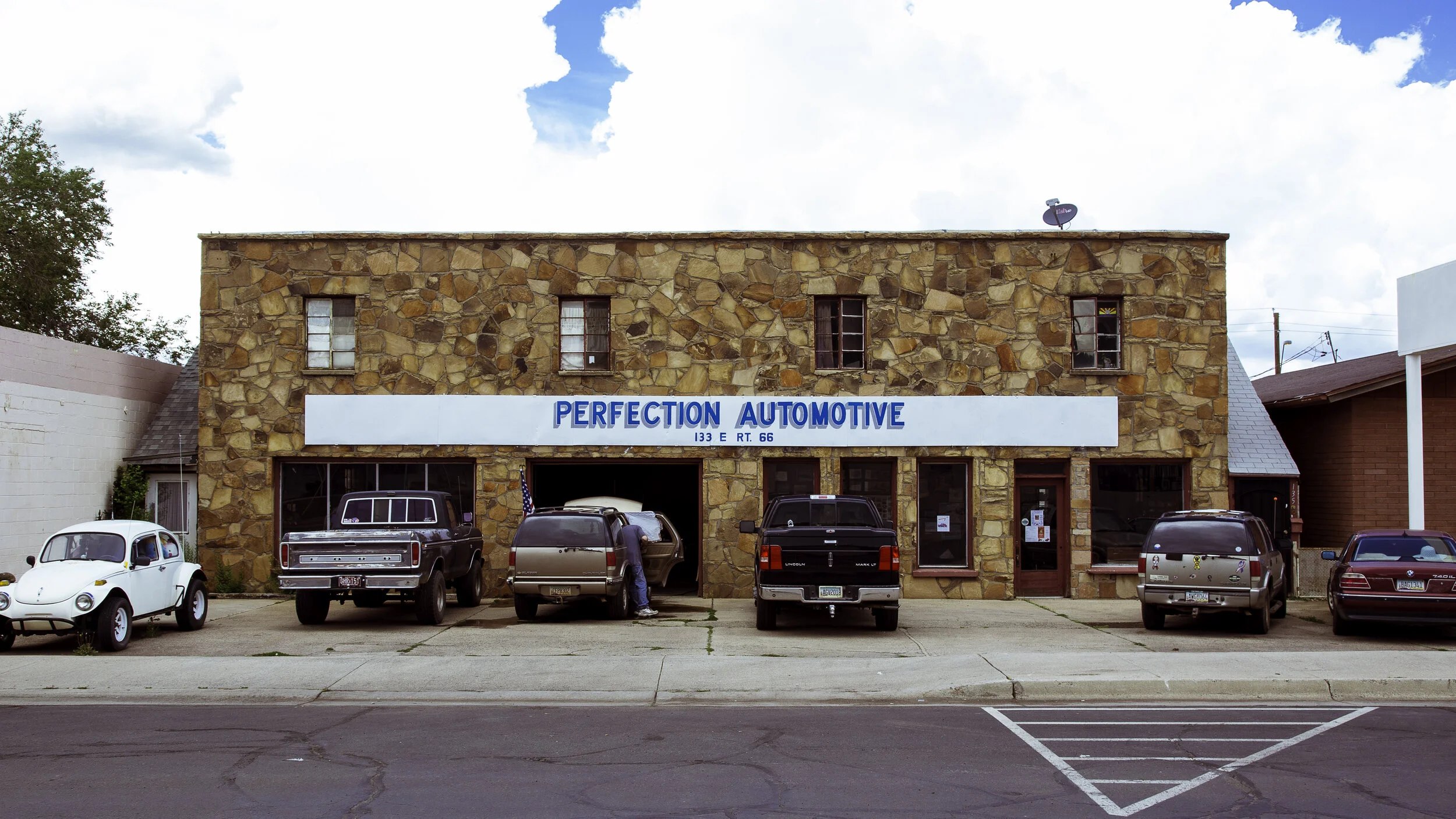A two-story stone building with a sign that reads "Perfection Automotive" and several parked cars in front, including a vintage white Volkswagen Beetle and a black pickup truck.