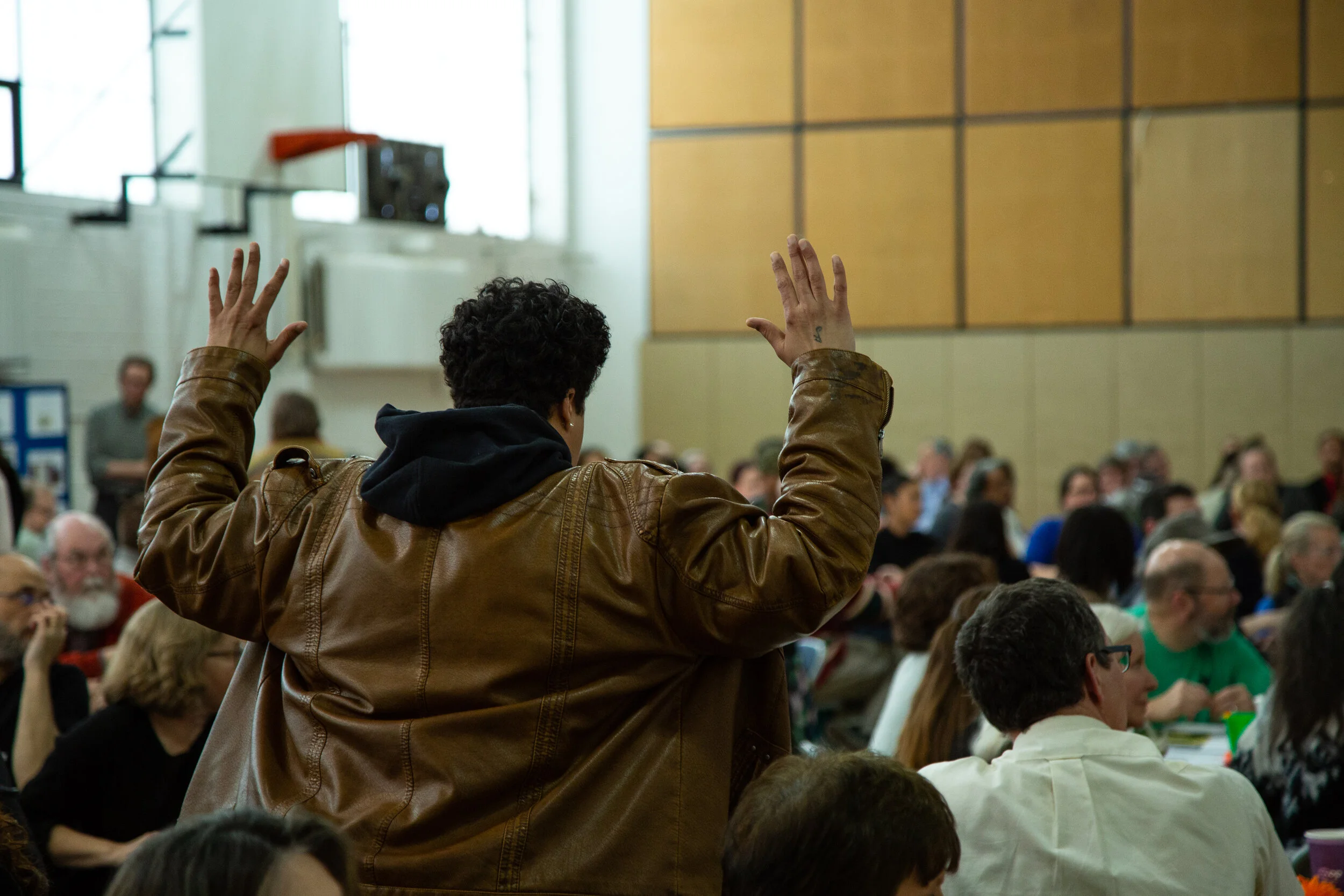 A person with short curly hair wearing a brown leather jacket and black hoodie raising their hands during a large indoor gathering or meeting.